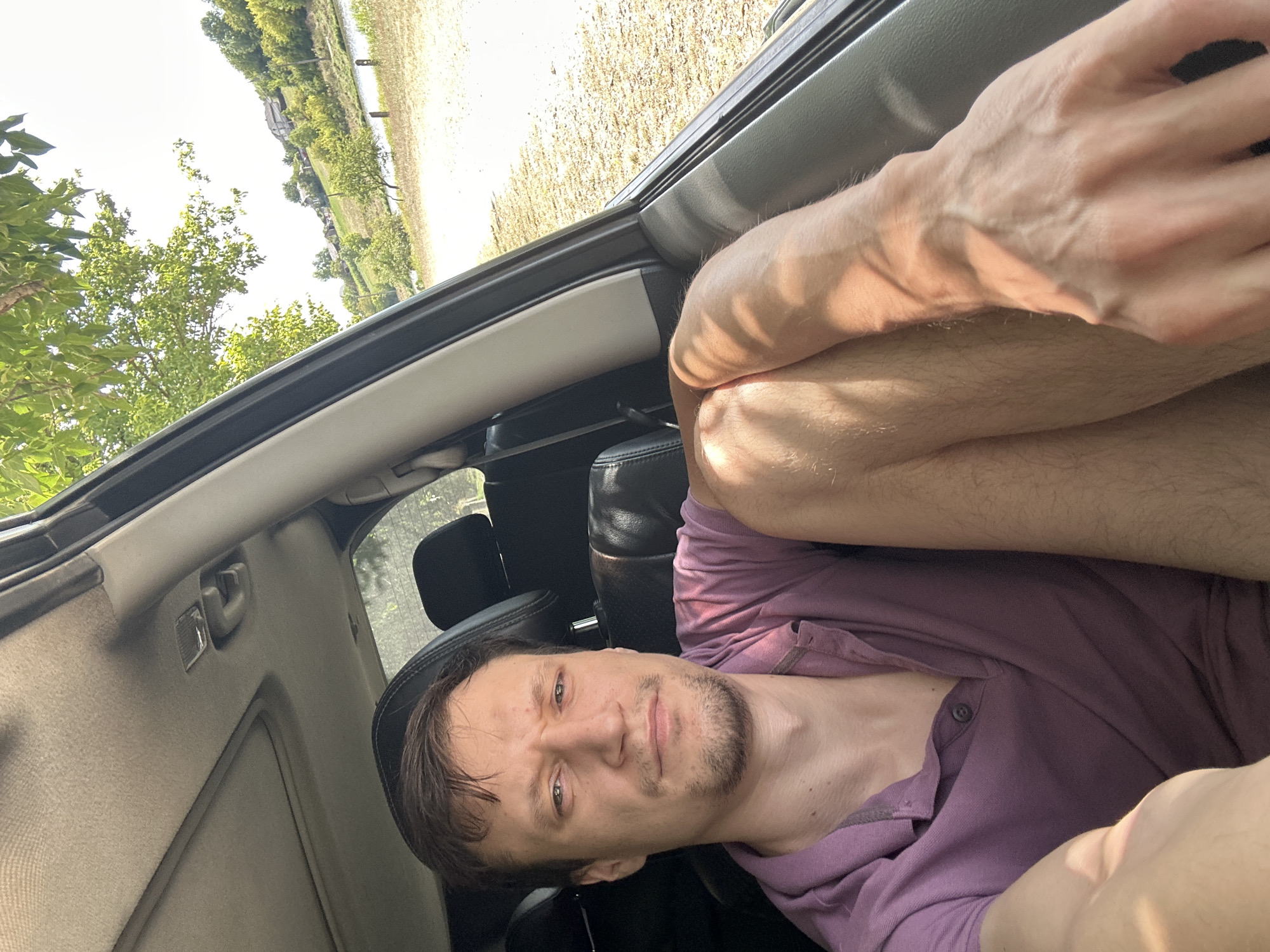 This is a selfie taken from inside a car, showing a man with short brown hair and a light beard sitting in the driver's seat. He is wearing a purple polo shirt. The man is looking directly at the camera with a neutral expression. His left arm is resting on his leg, with his hand in the foreground. Sunlight streams in through the open car window, illuminating his arm and hand. Outside the car, a landscape is visible, featuring a body of water, trees, and a few houses on a hill. The location is Makiv, Ukraine.