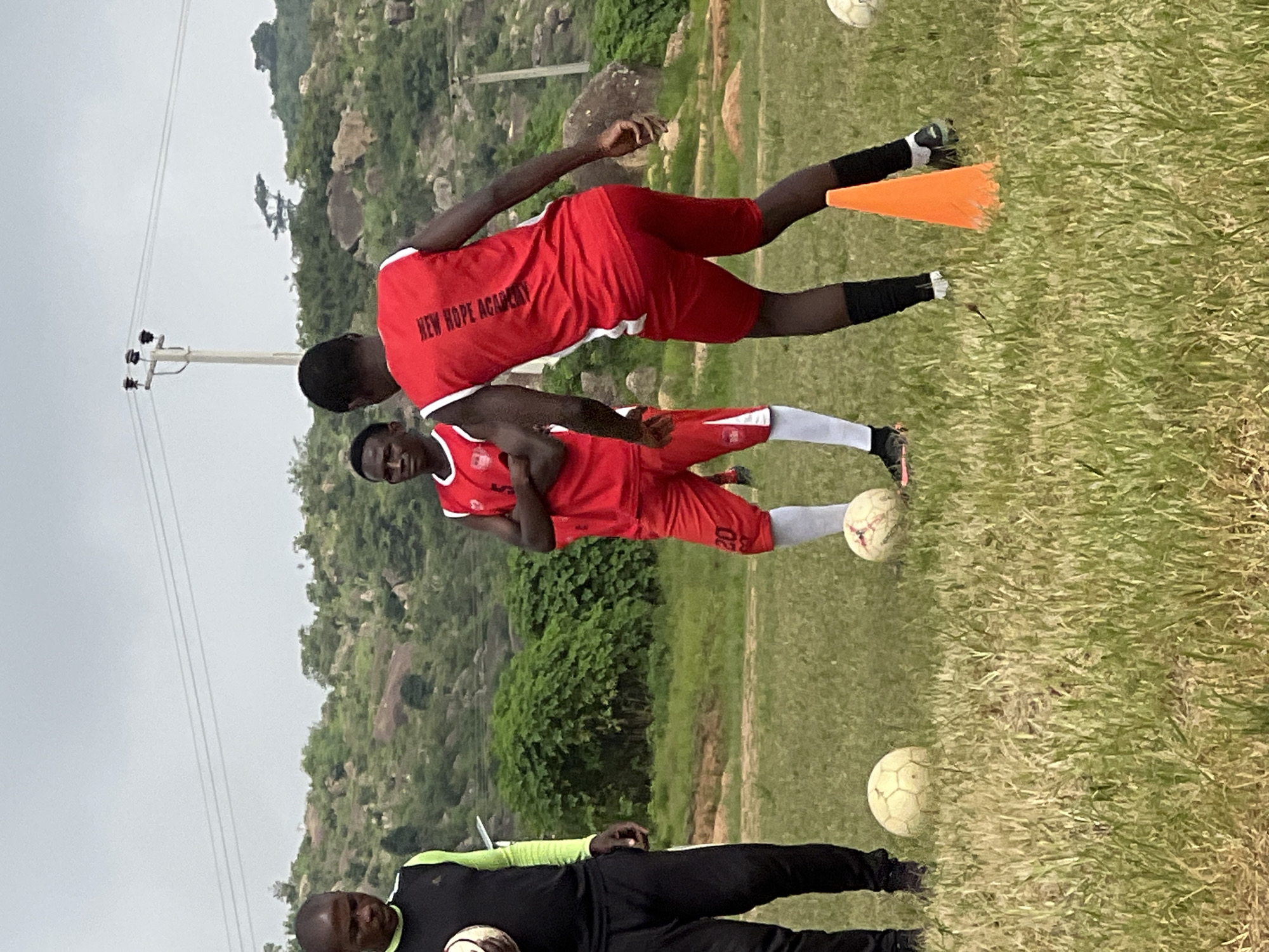 In Jos, Nigeria, on a grassy field under an overcast sky, three young men are engaged in a soccer training session. The man on the left, an adult, wears a black vest with lime green trim over a long-sleeved shirt and black track pants, holding a soccer ball. To his right, a young man in a red jersey with white trim and the number 5, red shorts, and white socks stands with his arms crossed. In the foreground and to the right, another young man, also in a red jersey with white trim and the number 5 visible on the back, wears red shorts, black socks, and cleats. He is positioned in a dynamic stance, as if in motion, with an orange cone placed near his feet. Two soccer balls are visible on the ground in the foreground. The background features lush green bushes and rocky hills, with a utility pole and power lines visible in the distance, indicative of an outdoor training area.