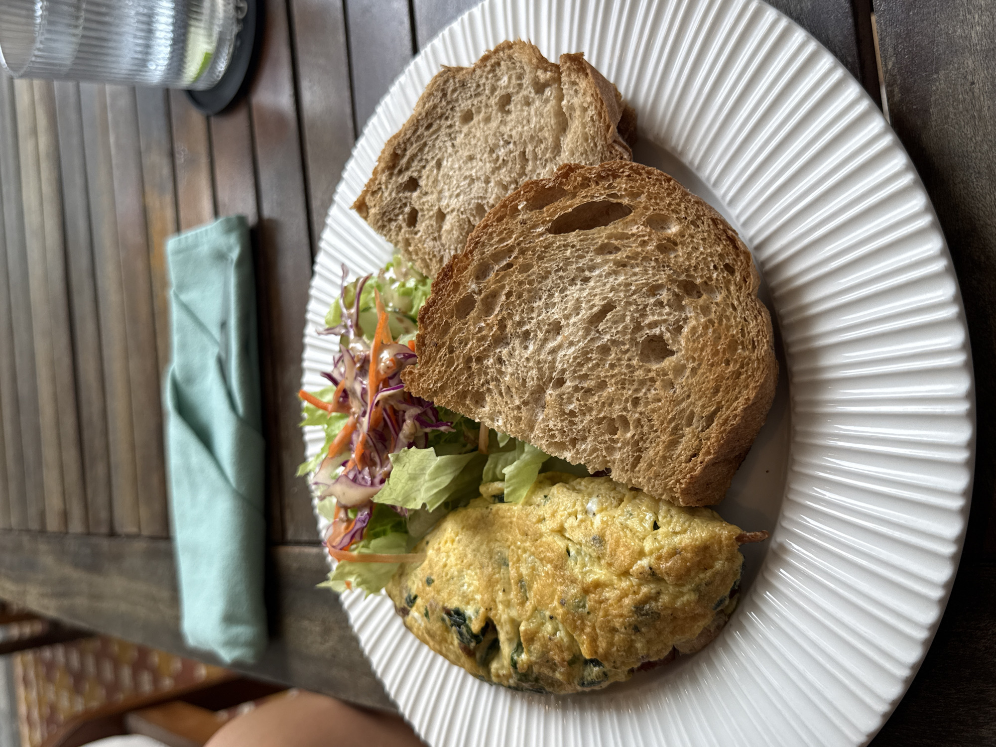 A close-up shot shows a plate on a wooden surface with a partially assembled sandwich. The sandwich consists of two slices of whole wheat bread, an omelet with visible green herbs (likely spinach), and a small salad of lettuce, shredded red cabbage, and carrots. The plate is white with a ridged design. A folded light-blue cloth napkin is to the left of the plate. In the upper left corner, there's a partial view of a glass with a lime wedge. The photo appears to be taken outdoors on a wooden table. The image provides no information to infer the location in Đà Nẵng, Vietnam.