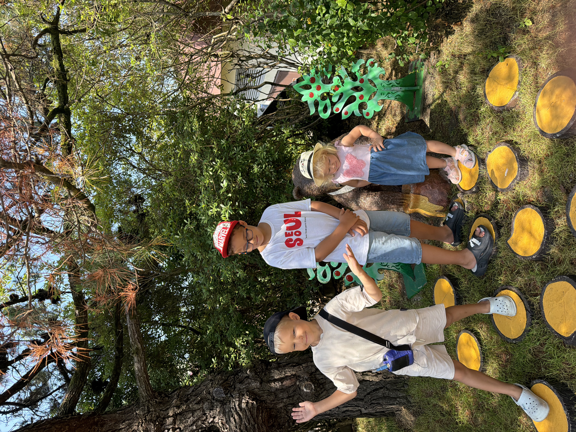 In an outdoor setting that appears to be a park or garden area in Sochi, Russia, three children are present. A young boy with blond hair, wearing a black baseball cap, a light-colored t-shirt, and light shorts, stands on a yellow stepping stone. He has a blue device attached to his side with a black strap. To his right stands another boy wearing a red cap with "MARVEL" on it, glasses, a white t-shirt with the word "SOUL" printed on it, and blue denim shorts. He is looking down. To the right of the second boy is a young girl wearing a straw hat, a pink and white striped top with a butterfly design, and a blue denim skirt. She is standing with her hands on her hips. Behind the children are decorative elements including what appears to be a bear statue, a green metallic cutout of a tree with red circles, and dense green foliage. The ground is covered in grass and yellow stepping stones, suggesting an interactive play area. The overall scene is illuminated by natural daylight, indicating it is daytime.