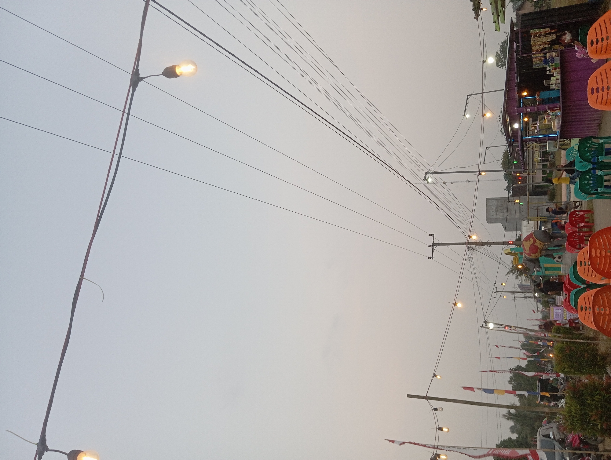 The image depicts an outdoor scene at dusk, likely a marketplace or roadside eatery in Way Dente, Indonesia. Overhead, a complex web of electrical wires stretches across the frame, with several bare incandescent bulbs strung along them, providing a warm, dim illumination. The sky is a pale, hazy gray, suggesting twilight or overcast conditions.
In the foreground, rows of brightly colored plastic chairs, predominantly orange and green, are arranged as if awaiting patrons. Behind the chairs, the environment opens up to reveal more of the setting. To the left, several flags with red and white patterns, possibly Indonesian flags, are mounted on poles, interspersed with other colorful flags. Vehicles, including a silver car and a motorcycle, are visible parked along the left side of the scene.
Further back, a row of small stalls or vendors can be seen. One prominent stall is a purple structure with an open front, displaying various items on shelves, suggesting it might be selling drinks or snacks. In front of this stall, people are present, some appearing to be customers or staff. To the right of the purple stall, a large, decorative elephant figure, adorned with red and yellow fabric, stands prominently. Behind the stalls, more structures and trees are visible, fading into the background. The overall atmosphere is that of a casual, open-air gathering place, possibly a night market or a local spot for food and socializing, illuminated by strings of lights as daylight fades.