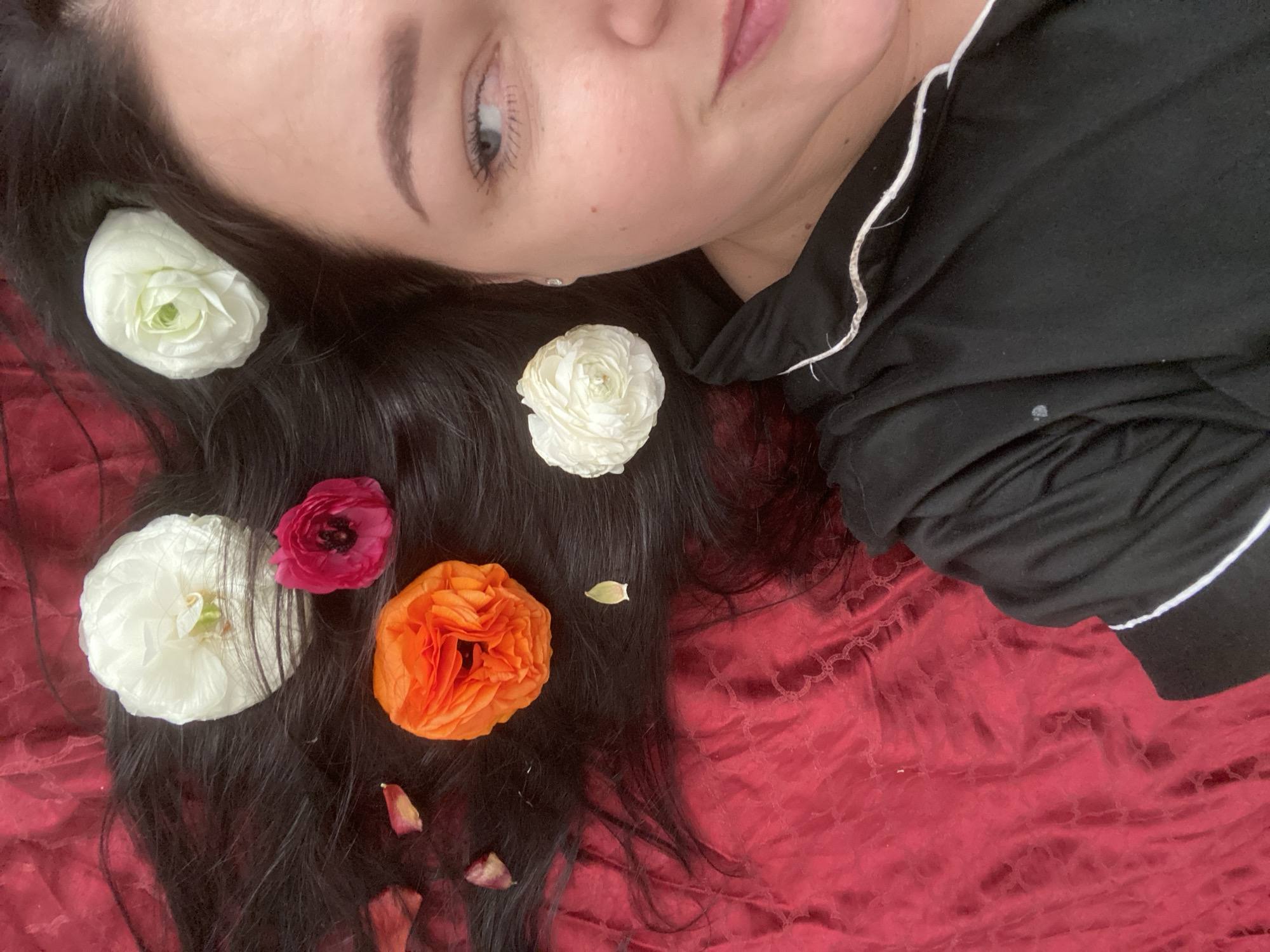A close-up, high-angle shot captures a woman lying down, viewed from her nose to the top of her head, with her dark hair spread out. Her left eye is open, gazing gently upwards, conveying a calm and serene expression. She is wearing a black top with white piping visible on the shoulder.
Her flowing, dark hair is adorned with several vibrant ranunculus flowers: three white blooms, one striking orange flower, and one deep pink one. A few scattered dried flower petals add to the organic arrangement around her hair. The woman is resting on a rich, textured red fabric, possibly velvet, which serves as the background, enhancing the visual warmth and depth of the scene.
The image portrays a peaceful and artistic moment, emphasizing natural beauty and relaxation. There is no visible text. This scene is set in Sochi, Russia.