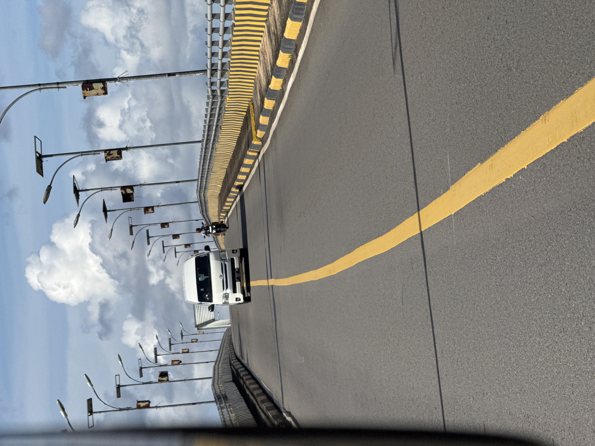 The image depicts a view from a vehicle traveling on a bridge in Tembeling, Indonesia. A white van is seen in the distance, followed by a person on a motorcycle. The bridge has a yellow and black striped barrier on the right side, with streetlights lining the left side. The sky is cloudy with patches of blue. The road is paved and has a yellow lane marker.