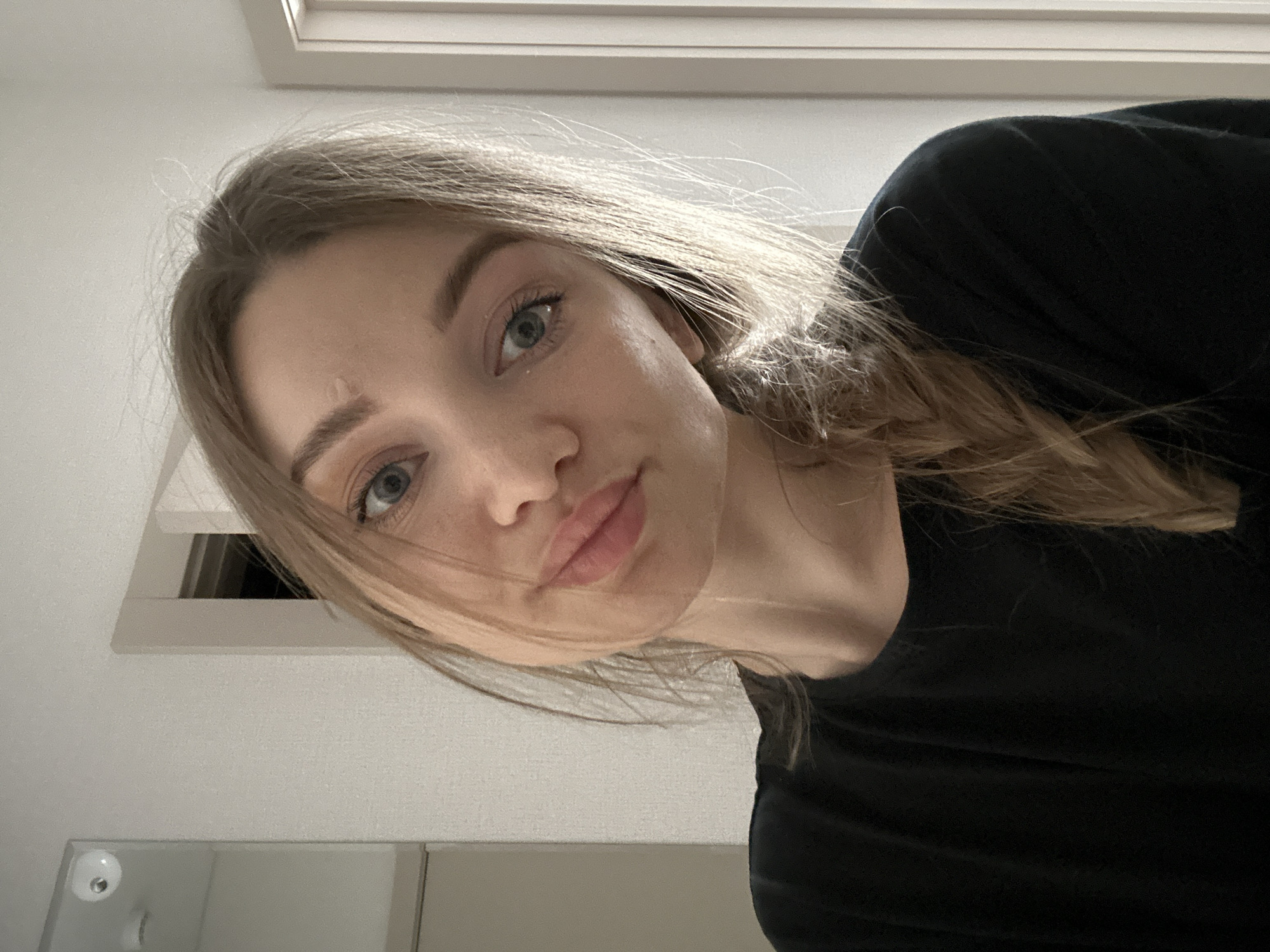 A close-up, high-angle view of a fair-skinned young woman with light brown hair and blue eyes. Her hair is partially braided and falls to the side. She is wearing a black top. A small scar is visible on her forehead. The background includes a white wall with a window or skylight near the top of the frame, and a mirror or reflective surface near the bottom. There is no contextual information to suggest this photo was taken in Kazan, Russia, or any other specific location.