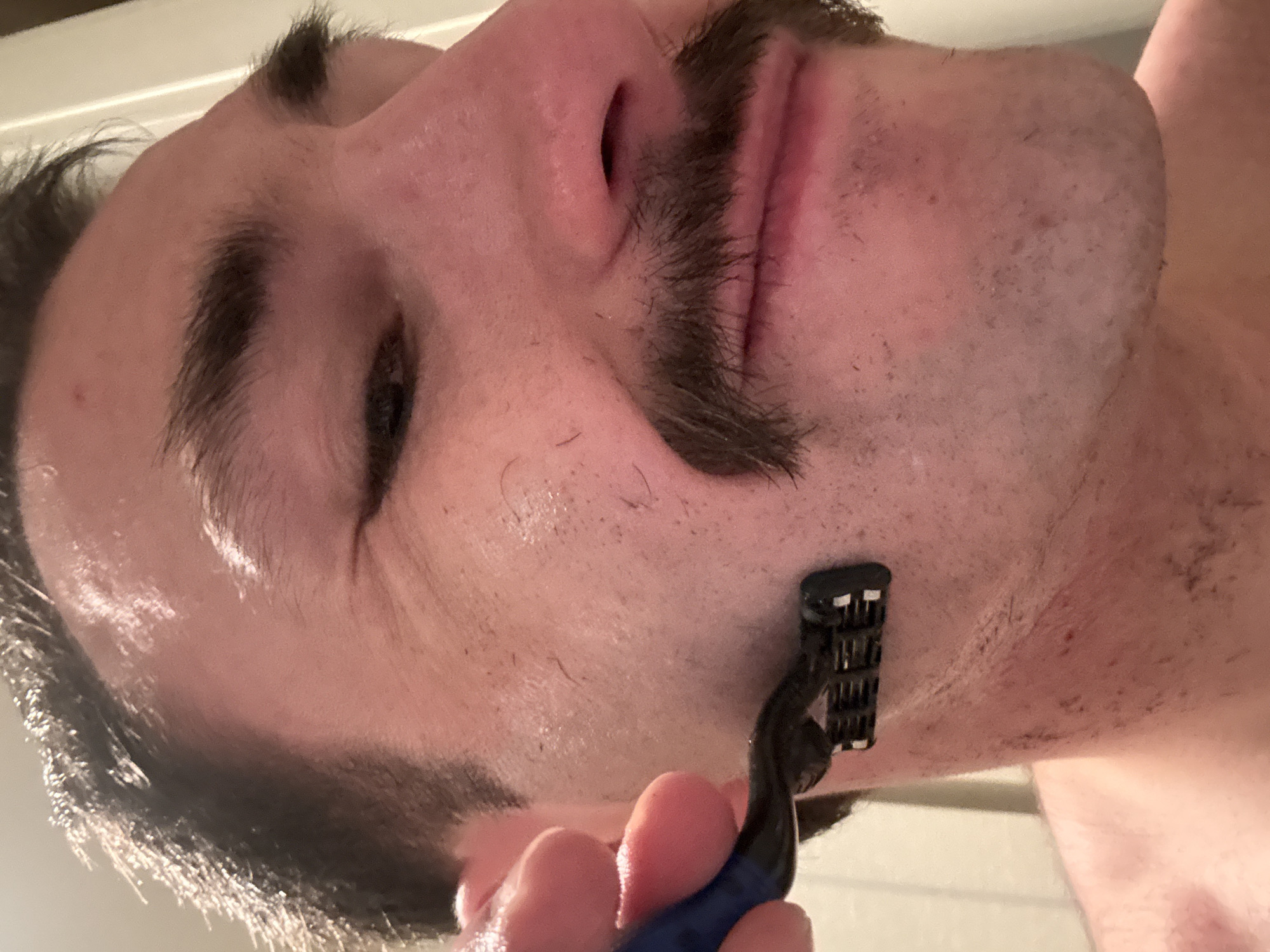 An upward-angled close-up shows a man in the process of shaving his face. The image is rotated sideways, depicting the man's head from the chin upwards. He has dark hair, thick eyebrows, and a prominent dark mustache. His eyes are squinted or mostly closed, and there's a slight grimace on his face, suggesting concentration or discomfort during shaving.
He is holding a multi-blade cartridge razor with a dark head and a blue handle against his right cheek/jawline. The razor appears to be actively shaving this area, and the skin around it looks wet, possibly from water or shaving cream. Visible stubble and small hair trimmings are scattered on his skin, particularly on the unshaved areas and where the razor has passed. Some patches of redness or irritation are also present on the freshly shaved skin. The man's mustache is well-defined and maintained, indicating it is being kept while the rest of his facial hair is being shaved. The scene appears to be indoors, likely in a bathroom. No text is visible in the image. The location context for this activity is Huixquilucan de Degollado, Mexico.