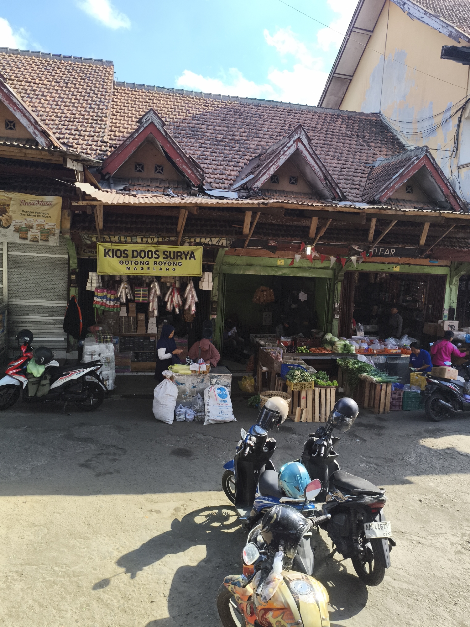 The image depicts a street scene in Magelang, Indonesia, featuring a market stall and several parked motorcycles. The stall, labeled "KIOS DOOS SURYA GOTONG ROYONG MAGELANG," displays various goods, including what appear to be spices or dried food items hanging from the ceiling and bags of groceries. In front of the stall, two women are visible, one tending to goods and the other in a conversation. Several motorcycles, some with helmets, are parked on the paved street in the foreground and midground. The architecture features traditional tiled roofs and wooden structures. The environment suggests a sunny day with some clouds in the sky.