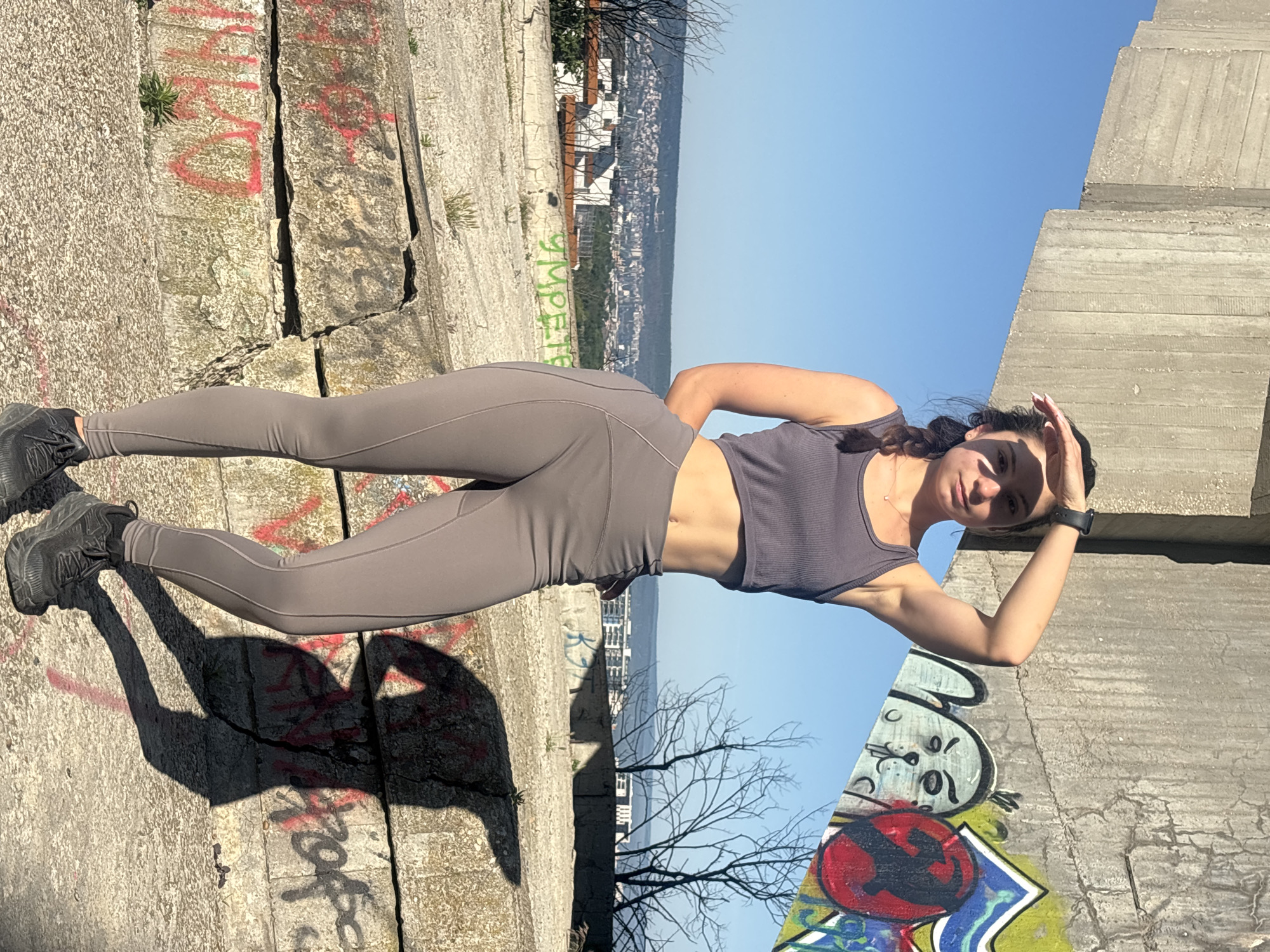 A young woman, wearing a grey athletic top and matching leggings with black sneakers, stands on a concrete structure with graffiti. She is shielding her eyes from the sun with her right hand. Behind her, a city skyline with buildings is visible under a clear blue sky. The location is likely an elevated point overlooking Varna, Bulgaria, given the presence of the city and the general appearance of the landscape. The graffiti on the wall and the abandoned concrete structure suggest an urban or semi-urban fringe setting.