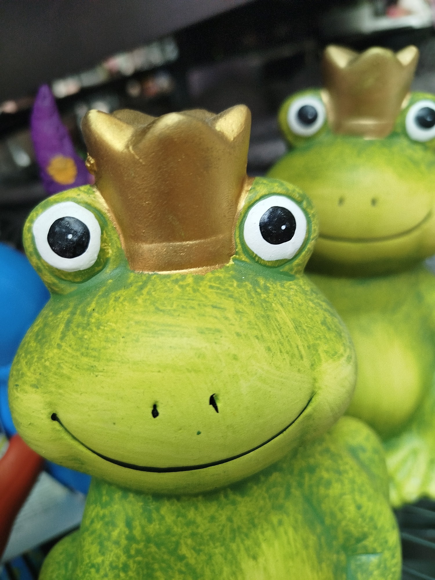 This is a close-up, eye-level, vertical shot of two decorative ceramic frogs, likely garden ornaments, positioned in the foreground. The frog on the left is in sharp focus, while the frog behind it is slightly out of focus. Both frogs are bright green with darker green speckling and wear golden crowns. They have large, round white eyes with black pupils. The frog in the foreground has a wide, smiling mouth with two small black dots representing nostrils. The background is dark and blurred, with hints of other objects, including what appears to be a purple gnome-like figure on the far left, and a red object to the left of the foreground frog. The scene is indoors, possibly in a shop or display area, and the lighting is soft. There is no indication of the specific location within Offenbach am Main, Germany.