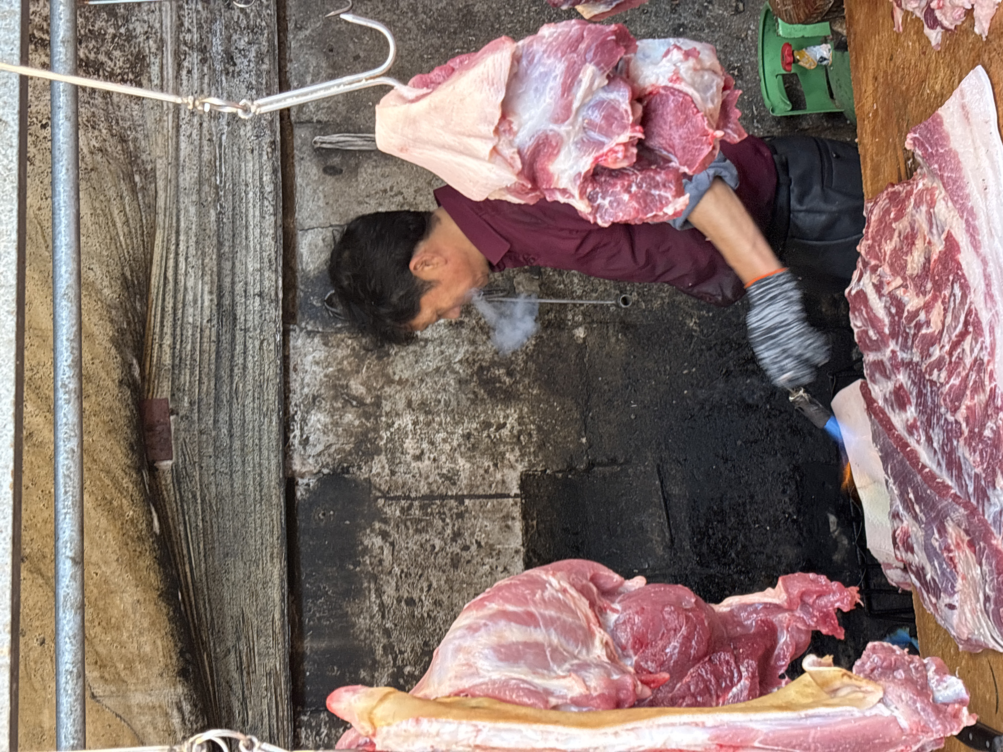 The image captures a scene at a meat stall in a market in Jiangjin, China, from an overhead perspective. The primary subject is a man with dark hair, dressed in a dark red long-sleeved shirt, dark trousers or apron, and a striped work glove on his right hand. A visible plume of white smoke or condensed breath emanates from his mouth, suggesting a cool or cold ambient temperature.
The man is actively engaged in handling large cuts of raw meat. A significant piece of meat, possibly pork, characterized by its pink muscle and white fat, hangs from a sturdy metal hook in the upper left. Another large, fleshy cut of raw meat rests on a wooden chopping block or counter surface to the right. Smaller pieces of red meat are arranged around the main cuts.
In the background, a green weighing scale is partially visible on the ground, indicating the process of selling meat by weight. The ground surface appears to be a worn mix of concrete and stained areas, typical of a busy market environment. A metal pole runs vertically along the left side of the frame, forming part of the stall's structure. The overall setting is utilitarian and focused on the butchering and sale of fresh meat. There is no visible text in the image.