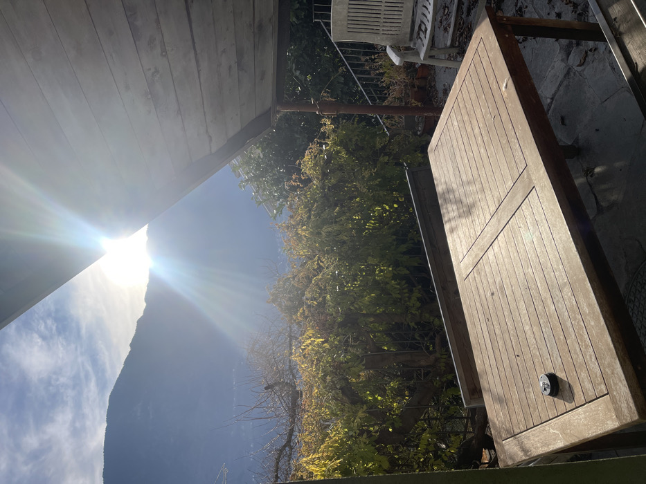 The image captures an outdoor patio or garden area, brightly lit by the sun, set against a backdrop of mountains. The scene appears to be taken from under a wooden-slatted roof structure, possibly a veranda or pergola, visible in the upper left.
In the foreground and mid-ground, a large, rectangular wooden table with a slatted top is prominently featured on the right. The table is weathered and has a small, dark, circular object with a lighter knob on its surface. Beyond the table, a white slatted garden bench is partially visible. The area behind the table and bench features lush green and yellow foliage, likely trees or large shrubs, with a thin pole or support structure extending vertically through them.
The background reveals a clear blue sky with some scattered white clouds, and a large mountain or hill rising sharply. Intense sunlight emanates from the upper left, creating a strong lens flare and illuminating the scene warmly. The low angle of the sun suggests either early morning or late afternoon. No people or specific activities are visible, and there is no discernible text. The overall atmosphere is peaceful and sun-drenched, typical of an outdoor setting in a location like Castione, Italy, with mountainous terrain.