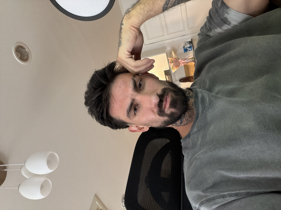 A male individual with dark hair, a full beard, and visible tattoos on his neck and left arm is seated in a black mesh office chair. He wears a grey, short-sleeved t-shirt and rests his left hand on his forehead, looking directly at the camera with a neutral expression. The scene is an indoor setting in Bangkok, Thailand. The background features a white ceiling with three white, egg-shaped pendant lights and a recessed downlight. An air conditioning vent is visible on the upper left. To the right, a white cabinet or shelf holds a bouquet of pink flowers in a clear wrapper, a small dark wooden object, and other indistinct items. A white paneled door is partially visible behind the cabinet. A large circular light reflector is also partially visible on the far right.
