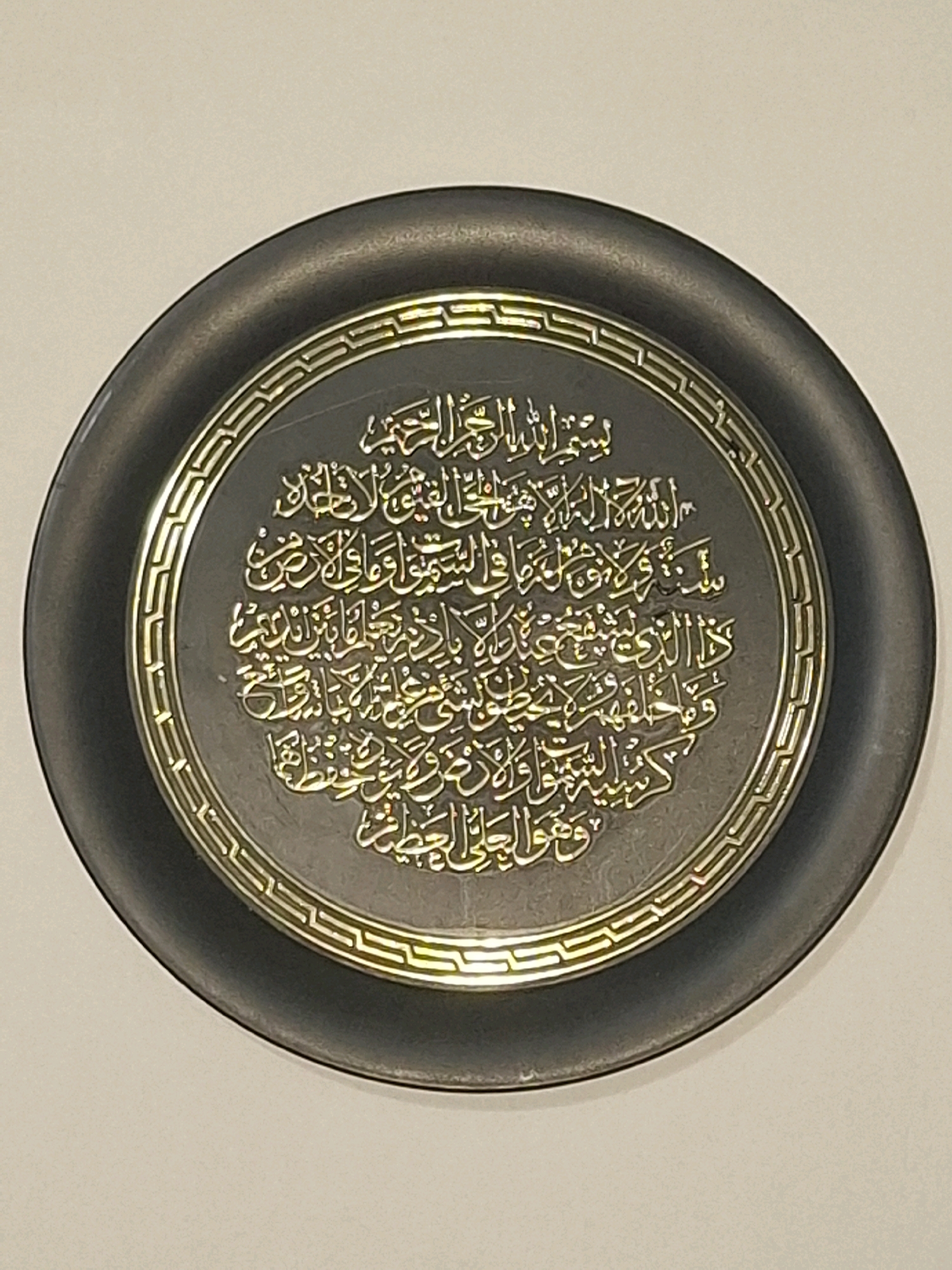The image shows a circular Islamic calligraphy plaque mounted on a wall. The plaque has a dark, possibly black, frame with a gold decorative border. The calligraphy itself is in gold and appears to be Arabic script, arranged in concentric circles. The text is highly stylized and ornamental. The plaque is centrally framed against a plain, light-colored background. There are no people or any recognizable landmarks of Almaty, Kazakhstan visible in the image.