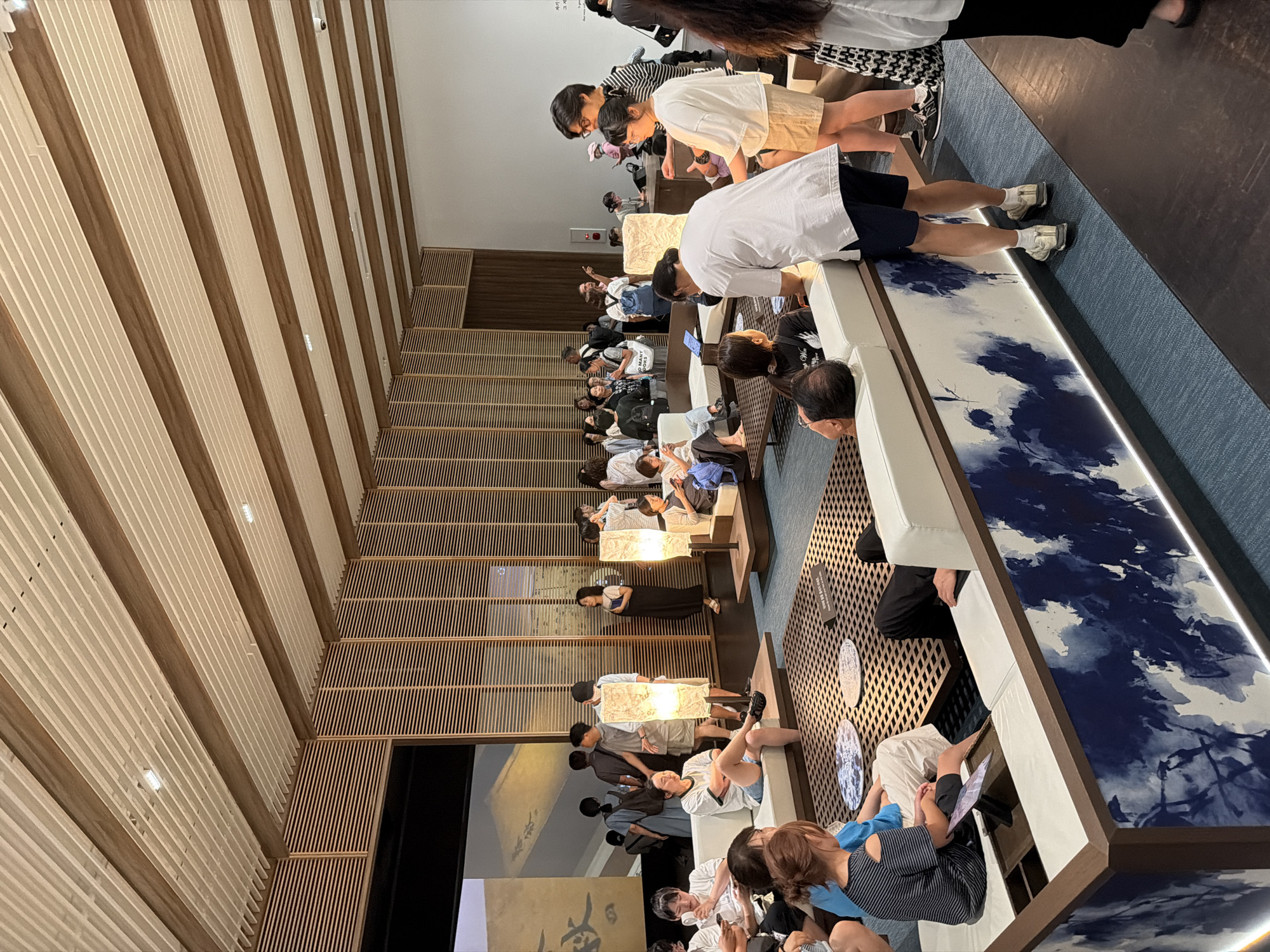 The image captures an indoor scene with several people casually interacting in a modern, artfully designed space. The environment features a prominent wooden slatted ceiling and walls, creating a warm and natural ambiance. Low-slung white modular sofas are arranged around a central, low-lying square coffee table with a patterned surface, suggesting a lounge or waiting area. Decorative floor lamps with a soft, diffused light are placed strategically.
The people in the image are dressed in casual attire. Some are seated on the sofas, looking at their phones or engaging in conversation. Others are standing, observing the surroundings or conversing with each other. There is a sense of relaxed informality.
In the background, a large screen displays an image with East Asian calligraphy. A wall with vertical wooden slats is a dominant architectural feature. The floor is covered with a textured blue carpet. The overall aesthetic suggests a contemporary cultural space, possibly an art gallery, museum, or a stylish public lounge area.
While the image doesn't explicitly state the location within Seoul, South Korea, the architectural style, the presence of art, and the demographic of the people present are consistent with a sophisticated cultural venue found in a major city like Seoul.