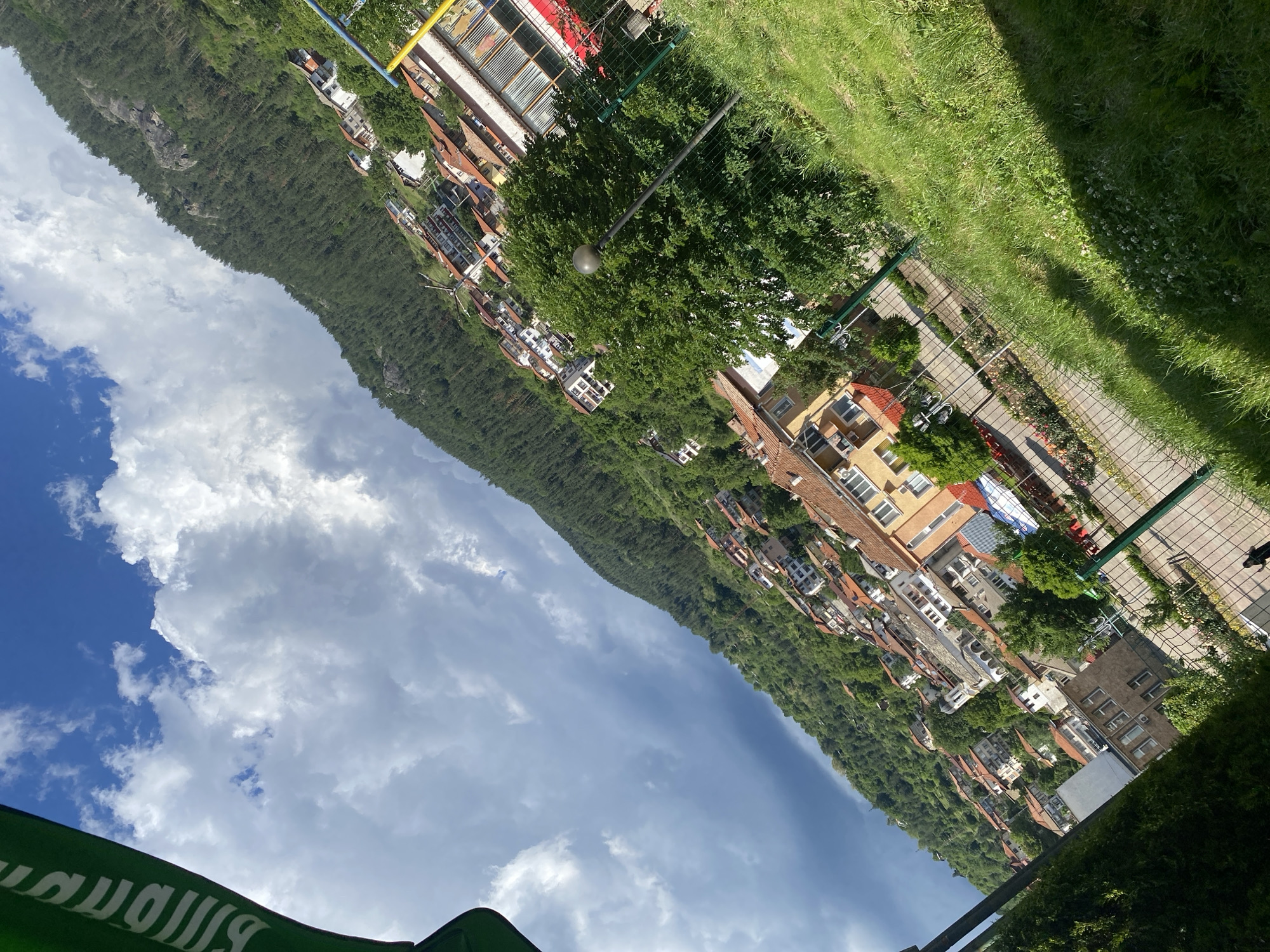 The image depicts a view of a hillside town in Smolyan, Bulgaria, on a cloudy day. The buildings are densely packed on the slope, with terracotta roofs and white or light-colored walls. Pine trees cover the upper parts of the hill. In the foreground, there is a grassy area with a fence and a lamppost. Part of a green banner with white lettering is visible in the upper left corner. The perspective is tilted, giving a sense of depth and height to the scene. No people are clearly discernible.