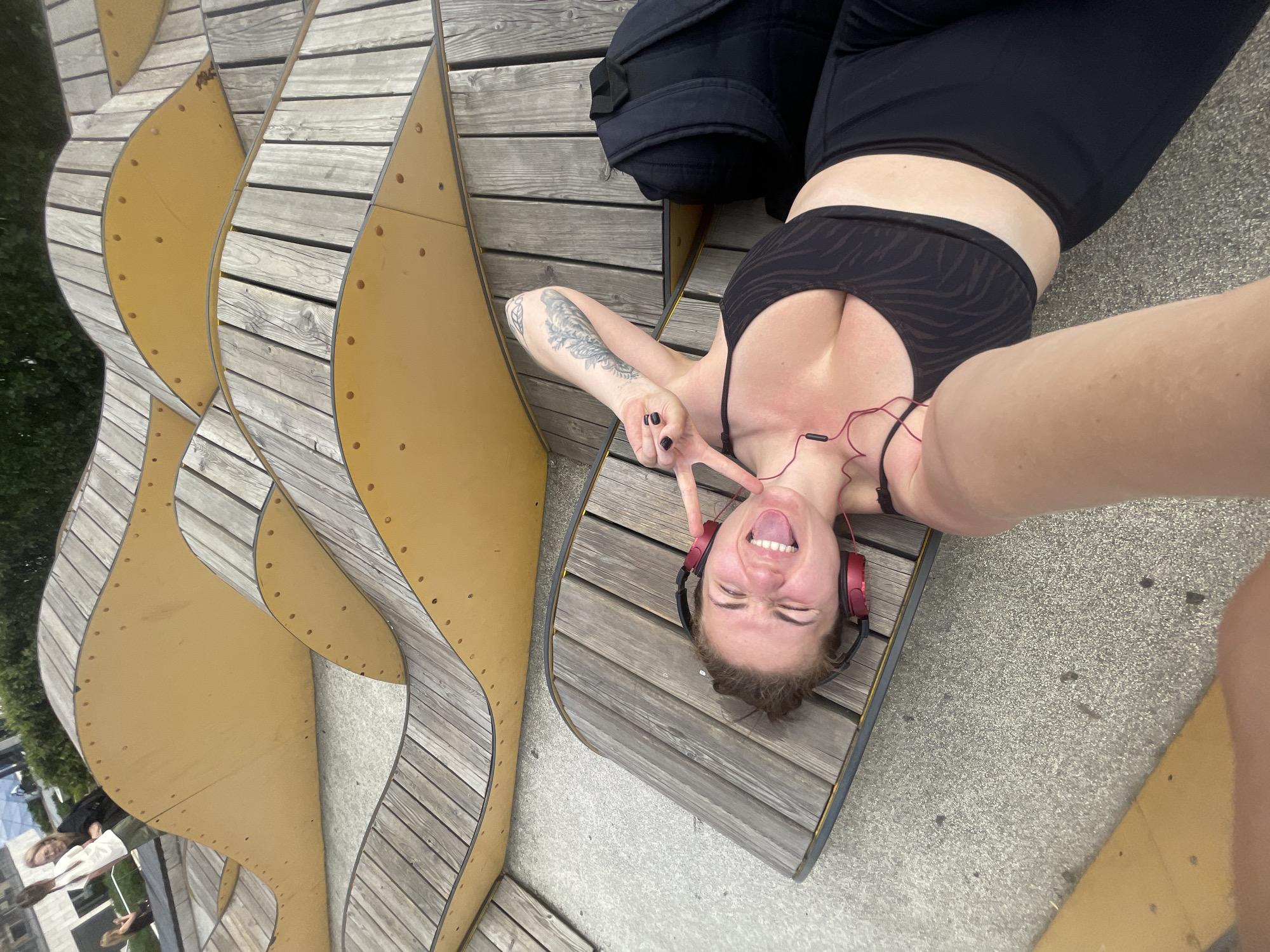 A woman is lying on her back on a wooden bench in an urban area, with a modern, wave-shaped structure behind her. She is wearing a black athletic top and black leggings, with red headphones around her ears. She is sticking her tongue out and making a peace sign with one hand. Her other arm is outstretched, suggesting she is taking a selfie. A black backpack is also on the bench near her head. The environment appears to be outdoors, possibly a park or public space within a city.