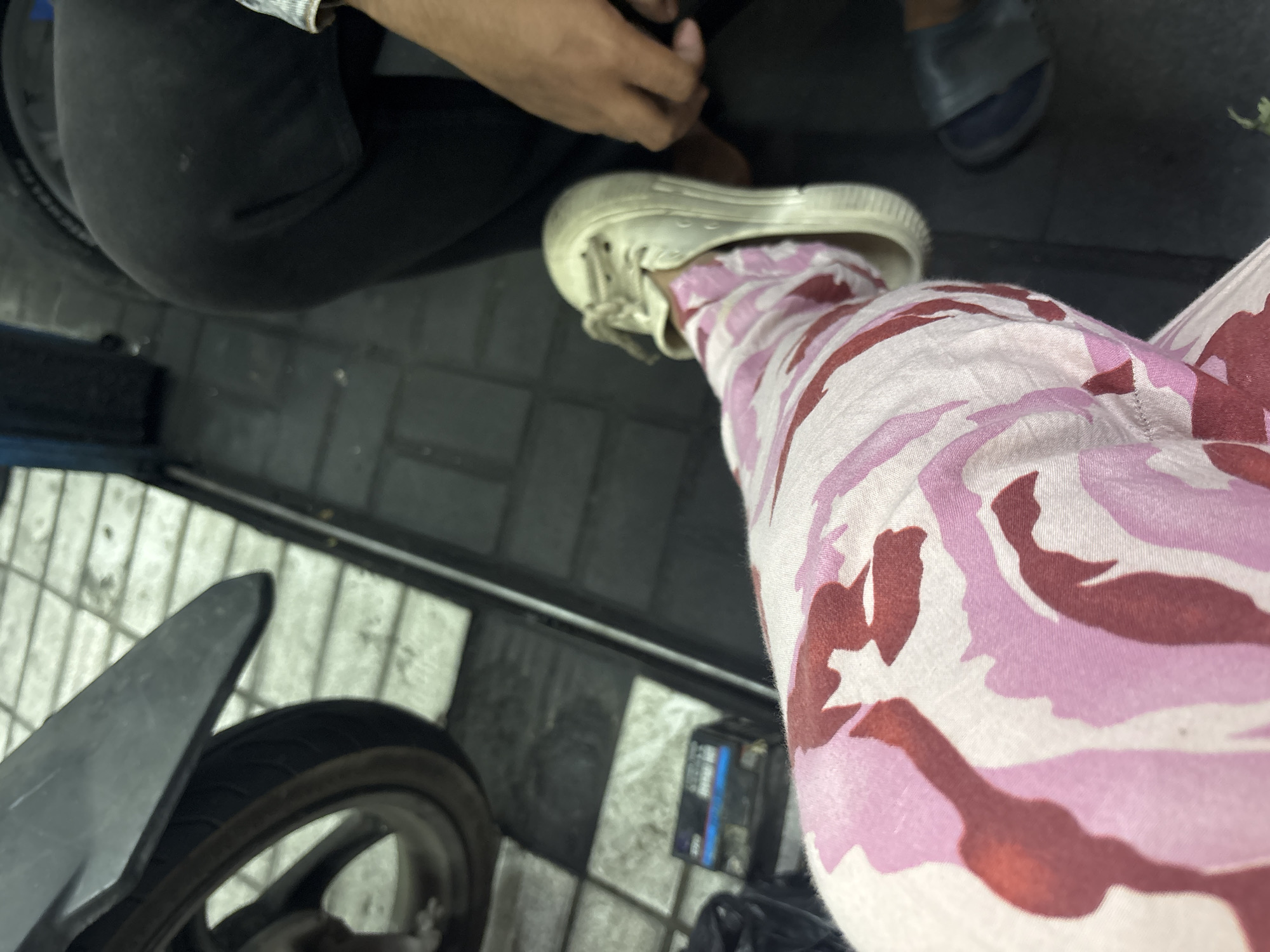 The image shows a close-up of a person's leg wearing pink and red camouflage pants and a white sneaker. The person is seated on a motorcycle, with the front tire and fender visible on the left side of the frame. In the background, another person is wearing black pants and a dark-colored shoe. The ground appears to be tiled with a checkered pattern of black and white squares. A black plastic object, possibly a battery, is visible on the ground near the motorcycle. The scene is outdoors, suggesting a street or parking area in North Jakarta, Indonesia.