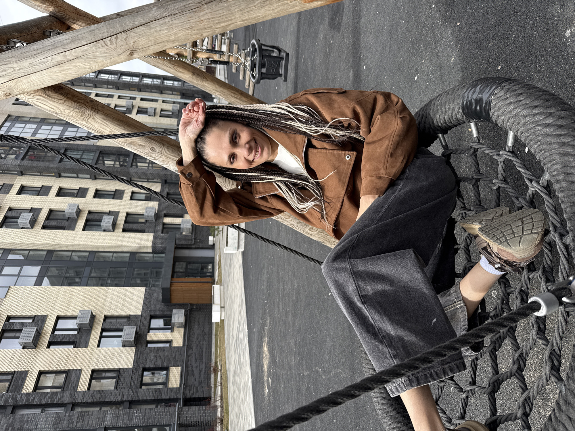 A smiling woman with braided hair, some of which is bleached white, wearing a brown jacket, white shirt, and gray jeans, is sitting in a black net swing in what appears to be a playground. She is holding her right hand up to her forehead.
In the background, there are several multi-story apartment buildings. The buildings are brick and have numerous windows with air conditioning units visible. The buildings are located in Rusanovo, Russia.
The playground has a black surface. Wooden support structures for the swing are visible. Another type of swing is partially visible in the background.