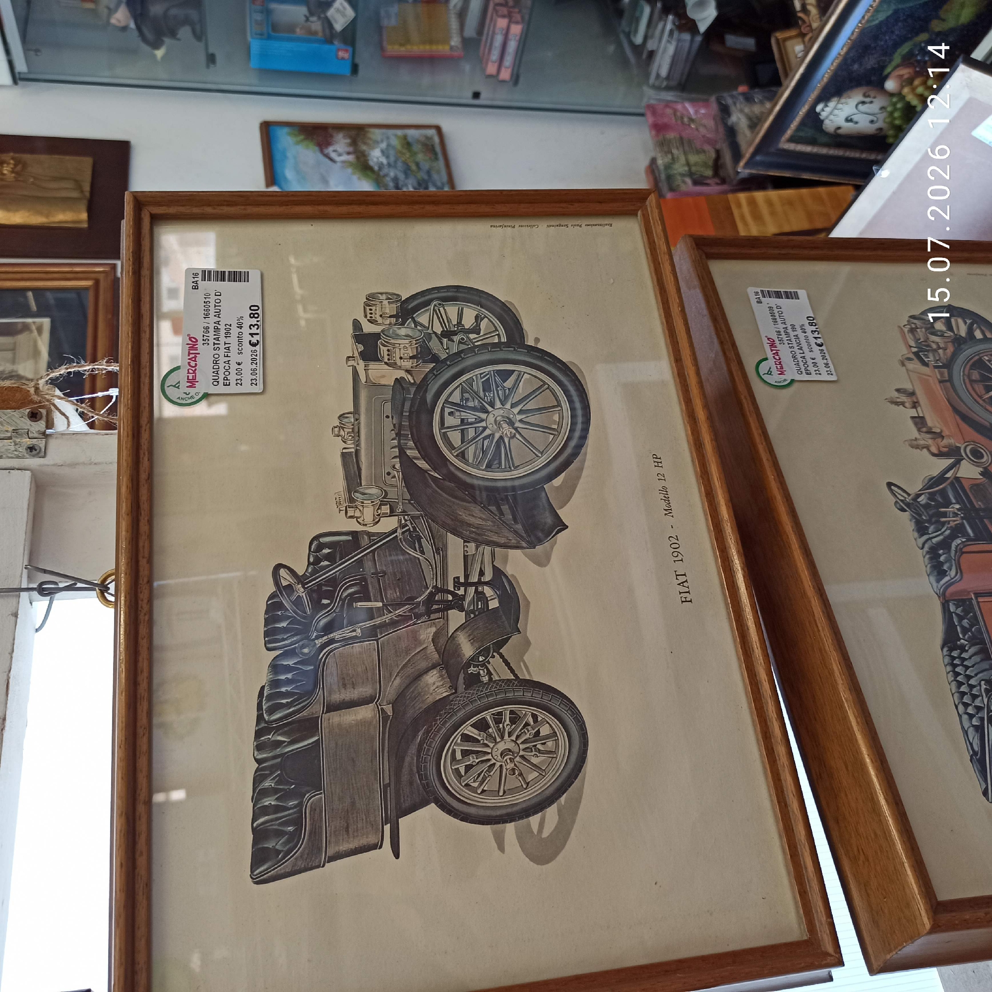 The image shows two framed prints of vintage cars. The top print features a detailed illustration of a FIAT 1902 - Modello 12 HP car, with a label on the right side indicating "MERCATINO" and sale details including a discounted price of €13.80. The second print, partially visible at the bottom, appears to be a similar vintage car print. Both prints are framed in light-colored wood. The background suggests an indoor setting, possibly a shop or antique store, with other items visible in the background including shelves with various objects, and a display cabinet with figurines. A watermark with the date "15.07.2026 12:14" is present in the bottom right corner. The location is identified as Molfetta, Italy.
