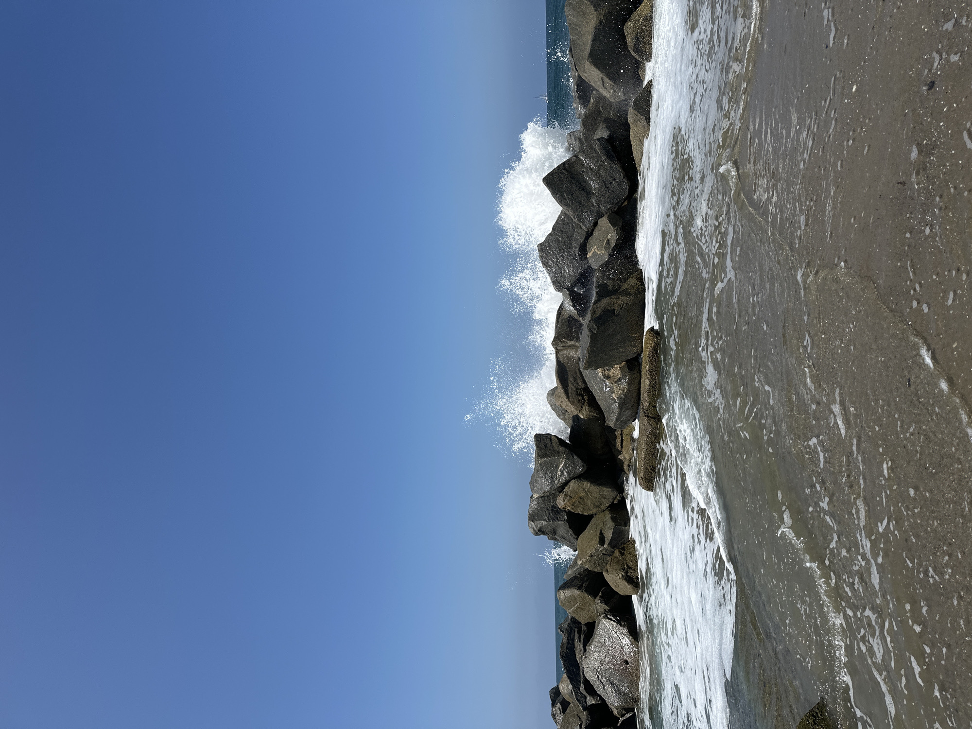 The image depicts a breakwater composed of large, dark rocks against a clear blue sky. Waves crash against the rocks, creating white spray and foam. The foreground shows the sandy shore with gentle waves lapping at the edge, creating a foamy surf. In the distance, the ocean stretches to the horizon, with a faint silhouette of a sailboat visible. The scene suggests a sunny day at a coastal location in Los Angeles, USA, likely a beach area with a man-made structure to control wave action. There are no people visible in the image.