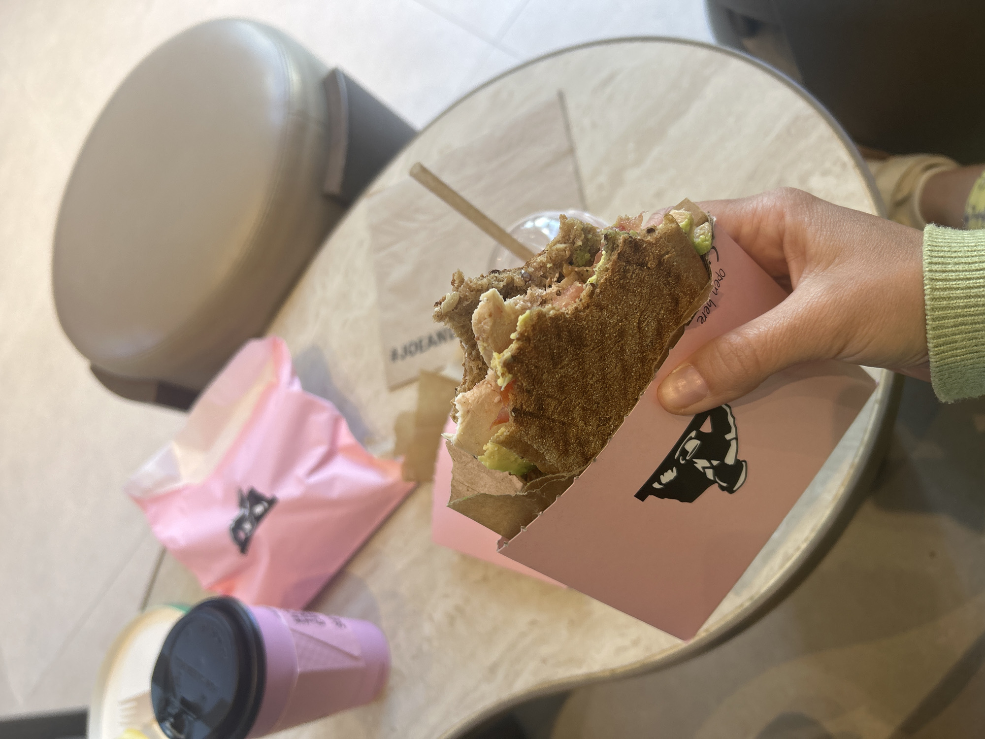 A medium overhead shot depicts a half-eaten sandwich in a pink paper container, held in a person's hand, on a light-colored round table. A light brown upholstered chair is visible on the left, with a portion of a dark upholstered chair visible in the upper right. A small pink cup with a black lid and another pink bag are near the sandwich on the table. A wooden stick and paper napkin are also present on the table. The location is likely a cafe in Casablanca, Morocco, based on the food container with possible French or Arabic writing. The person is presumably eating the sandwich.