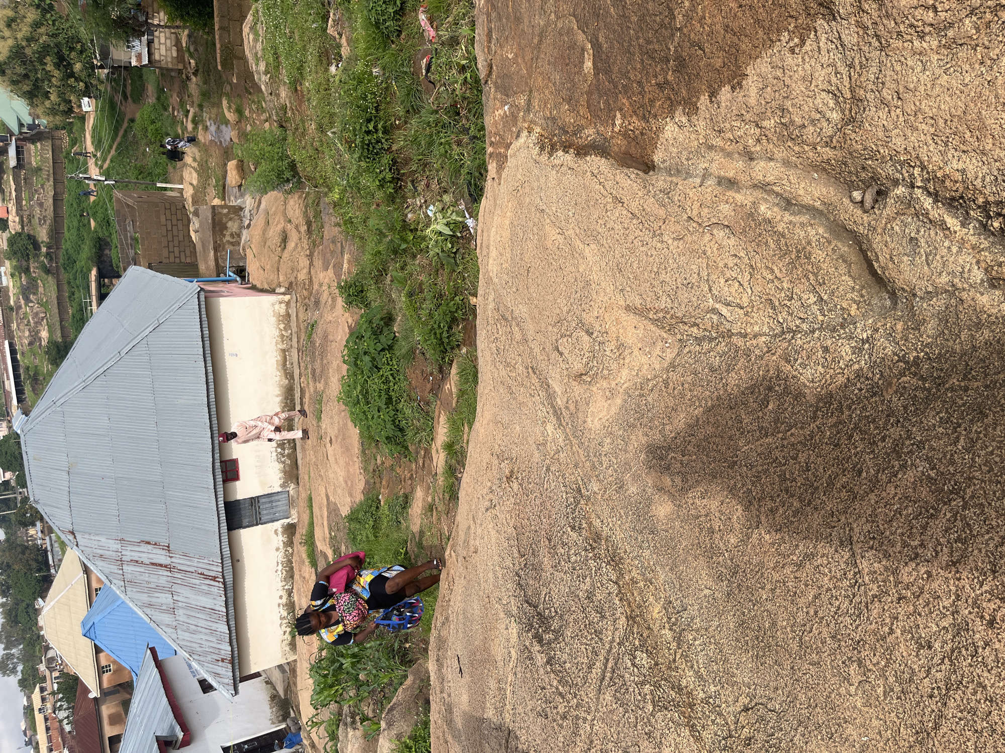 The image shows a scene in Jos, Nigeria, likely during the day with the sun casting shadows. The foreground is dominated by a large, rough-textured rock face. To the left, a settlement is visible, featuring various buildings with corrugated metal roofs and some with a lighter colored paint. A path or dirt road winds through the settlement, with a few scattered trees and vegetation.
In the middle ground, near the rock face and adjacent to a building, two individuals are present. One person appears to be a woman wearing a colorful patterned headscarf and a black dress, carrying a bag and looking down. Another individual, dressed in a light-colored traditional outfit, stands near the building, looking towards the left. Another person can be seen in the distance further up the hill.
The overall setting appears to be a mix of natural rocky terrain and a residential area. The presence of people suggests daily life activities are taking place. The light suggests it is daytime, and there are no clear indicators of the weather beyond the bright lighting. The buildings and vegetation are consistent with a rural or semi-urban environment in Nigeria. No text is visible in the image.