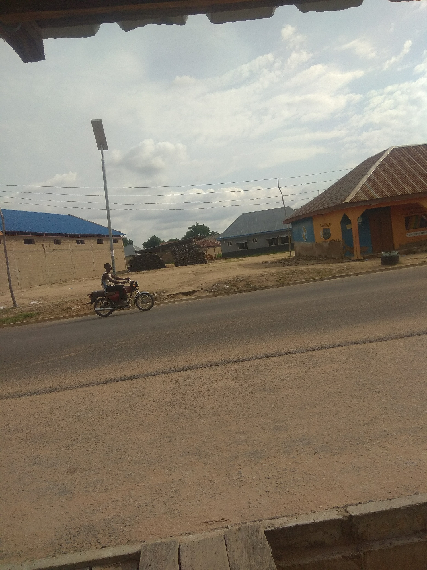 A lone motorcyclist rides a red motorcycle along a paved road in Jalingo, Nigeria. The motorcyclist is a Black male wearing a light-colored tank top and dark pants. The road is lined with buildings on the left and right. On the left, a large building with a blue roof and tan walls is visible. Further down the street, a building with a corrugated metal roof painted orange and yellow stands prominently. A tall, modern street lamp with a solar panel stands near the road. The sky is overcast with scattered clouds. The overall scene depicts a typical urban streetscape in Nigeria.