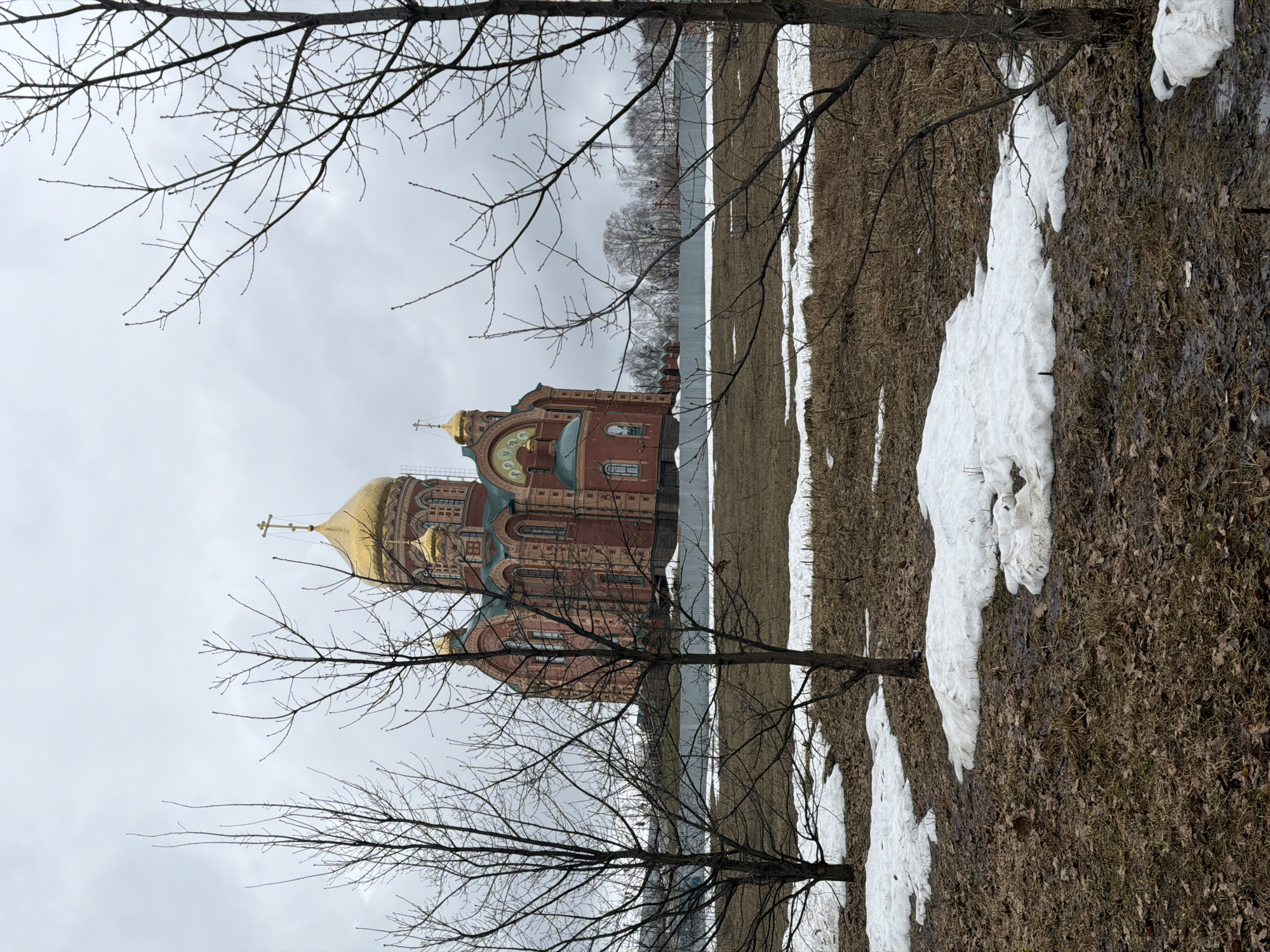 The image shows a partial view of the Church of St. Nicholas the Wonderworker in Berezniki, Russia, during a transitional season between winter and spring. The building is constructed of red brick with golden domes. The photograph is framed by bare tree branches, indicating a late winter or early spring setting. Patches of snow remain on the ground, which is otherwise covered in brown grass. The sky is overcast. The church is positioned on the edge of a body of water which is cut off by a wire fence.