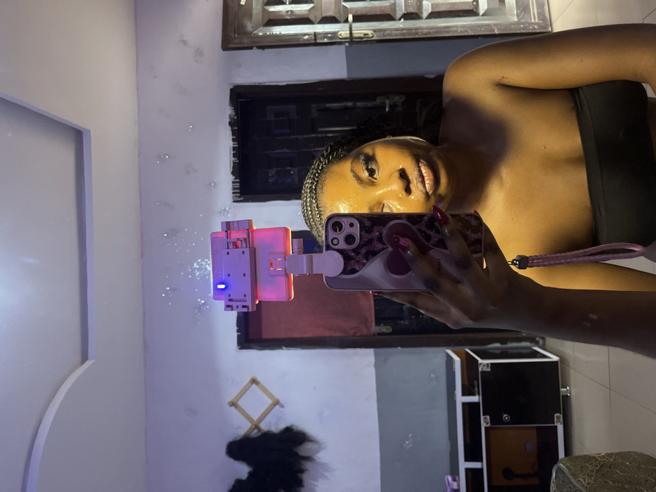 A young adult female is depicted taking a mirror selfie inside a room, presumably located in Gbongan, Nigeria. The subject, positioned centrally, has dark skin, braided dark hair, and wears a black strapless top. Her face exhibits a yellowish, possibly cosmetic, application and she has a nose piercing, with her lips slightly puckered. Her left arm is raised, holding an iPhone with a pink and white patterned case featuring a heart design. A pink braided wrist strap is attached to the phone. A clip-on ring light is affixed to the top of the phone, emitting a red glow downwards and displaying a blue indicator light.
The background, reflected in the mirror, reveals an interior room with a white ceiling featuring a decorative raised panel. The walls are light-colored, showing some texture or wear, with sparkling reflections visible, possibly from the ring light. To the left, a dark-colored wooden dresser or console table with lighter brown panels and silver legs is visible. Above it, on the wall, a light-colored wooden expanding rack holds dark, possibly fibrous, material. Two doorways are visible in the background: one partially obscured dark door frame to the left, and a more prominent dark door with a white frame and metal grating on the right side of the image. The floor appears to be tiled.