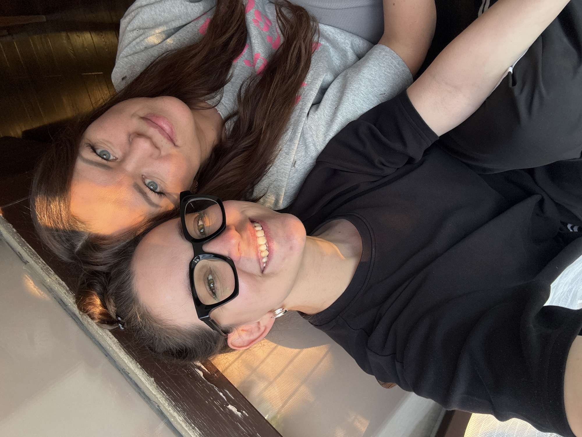 Two women are captured in a close-up, casual photograph, seemingly lying down and looking up towards the camera. The image is taken in what appears to be an indoor setting, likely in Chaikovsky, Russia, with warm, golden light illuminating their faces, suggesting late afternoon or early morning. Both women are smiling, conveying a sense of happiness and warmth.
The woman positioned on the left has long, dark brown hair flowing around her shoulders. She has clear blue eyes and a subtle, pleasant smile. She is wearing a light grey hooded sweatshirt with faint pink accents.
The woman on the right has her dark brown hair partially pulled back. She wears black-rimmed glasses, through which her green eyes are visible, and a prominent silver hoop earring on her left ear. She is smiling broadly, showing her teeth, and is dressed in a black long-sleeved shirt.
The background includes a section of dark wooden flooring or paneling, and a lighter, possibly tiled or window-adjacent, surface, further accented by the diagonal streaks of natural light. The overall mood of the image is relaxed and friendly. There is no visible text in the image.
