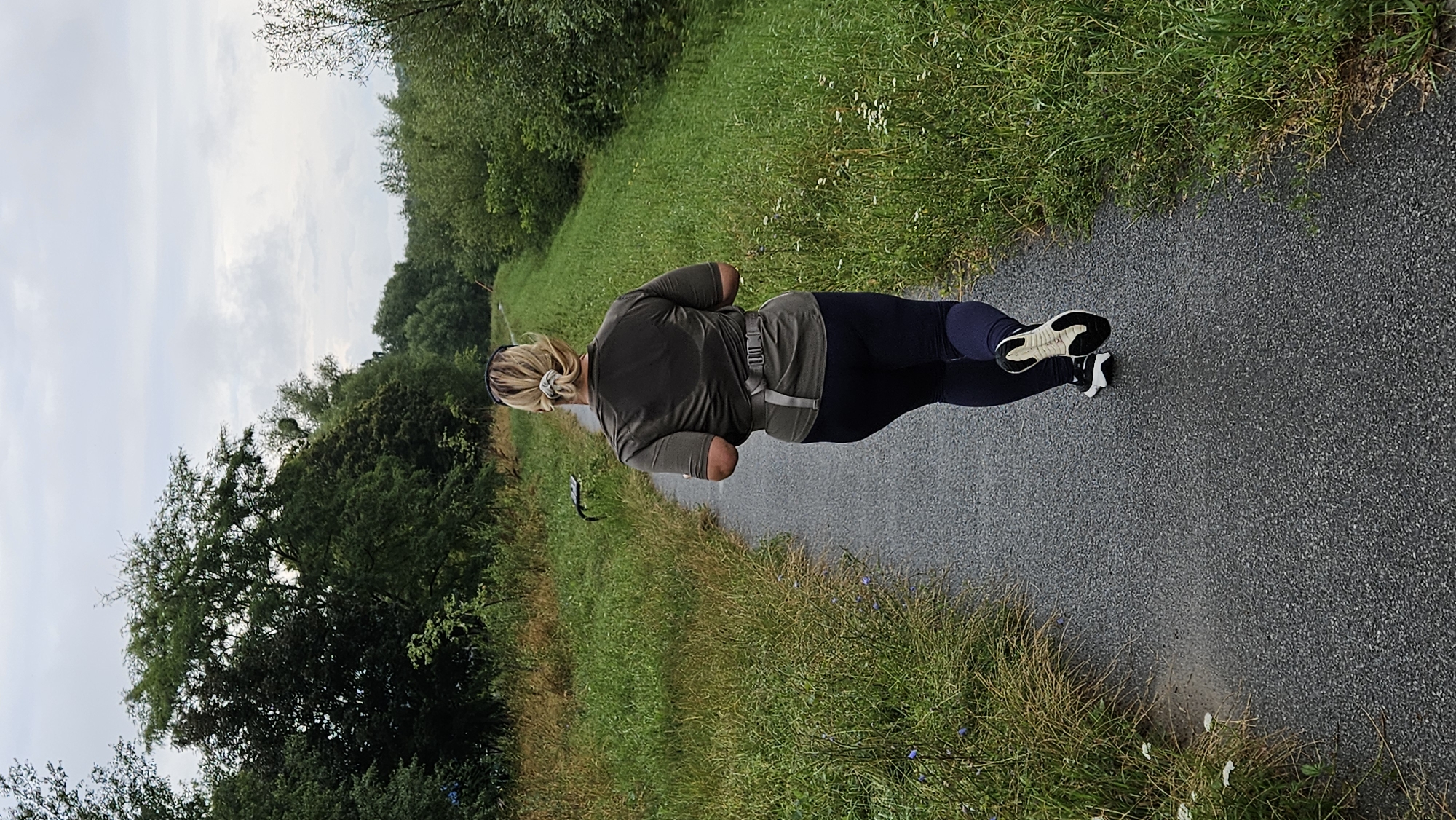 A rear view medium shot captures a woman running on a paved path flanked by lush green grass and trees. The woman, with blonde hair pulled into a bun, wears a dark olive-green t-shirt, dark leggings, and athletic shoes with white soles. A gray running belt is visible around her waist. The path curves slightly to the left as it recedes into the distance, bordered by a dense line of trees on the right and tall grass on the left. The sky is overcast with a few clouds. The scene is an outdoor park or nature trail. The presence of trees, grass, and a paved path suggests an urban park or a recreational area, potentially within or on the outskirts of Prešov, Slovakia.