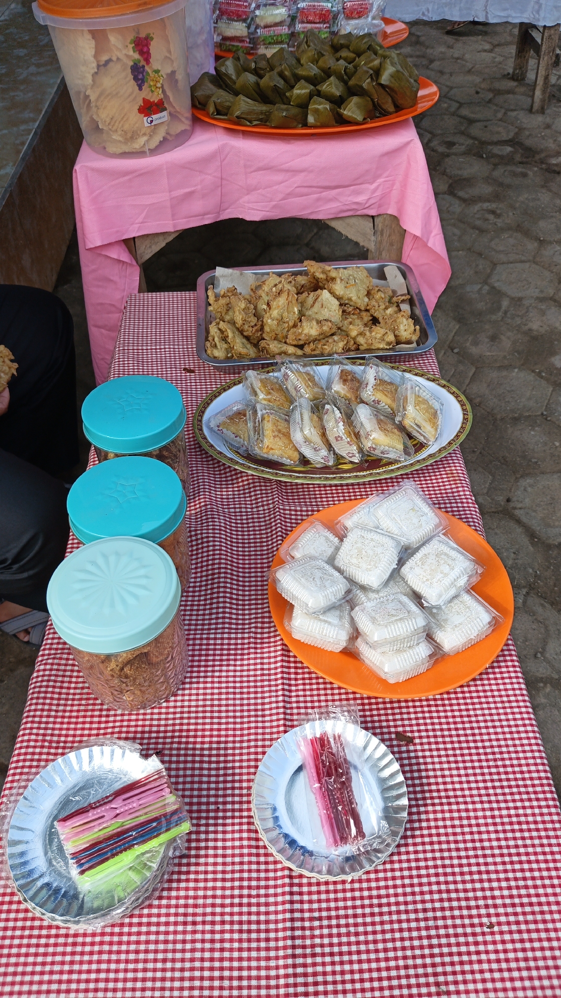 The image depicts an outdoor food stall, likely in Linggasirna, Indonesia, featuring a variety of snacks and sweets arranged on tables covered with a red and white checkered tablecloth. In the foreground, two silver paper plates are visible, one holding a stack of colorful plastic forks wrapped in cellophane, and the other holding several pink and red jelly sticks also in cellophane.
To the left of the plates are three clear plastic jars with teal lids, filled with what appears to be a dried, shredded snack. Further back, on a pink tablecloth draped over a wooden table, a large clear plastic container holds light-colored crackers. Beside it, an orange oval platter is filled with numerous green, leaf-wrapped parcels, likely a type of traditional sweet or savory snack. Behind this, a rectangular metal tray contains a generous portion of golden-brown fried items, possibly fritters or fried vegetables.
In the middle ground, an oval white platter holds multiple individually wrapped snacks, each appearing to be a layered pastry or cake. Adjacent to this, an orange round platter is laden with small, square, white confections, also individually packaged in clear plastic. The ground beneath the tables is paved with rough, irregular stones. A person's legs, wearing dark pants and sandals, are visible on the far left of the image, indicating a customer or vendor is present. The overall scene suggests a lively market or roadside stall environment.