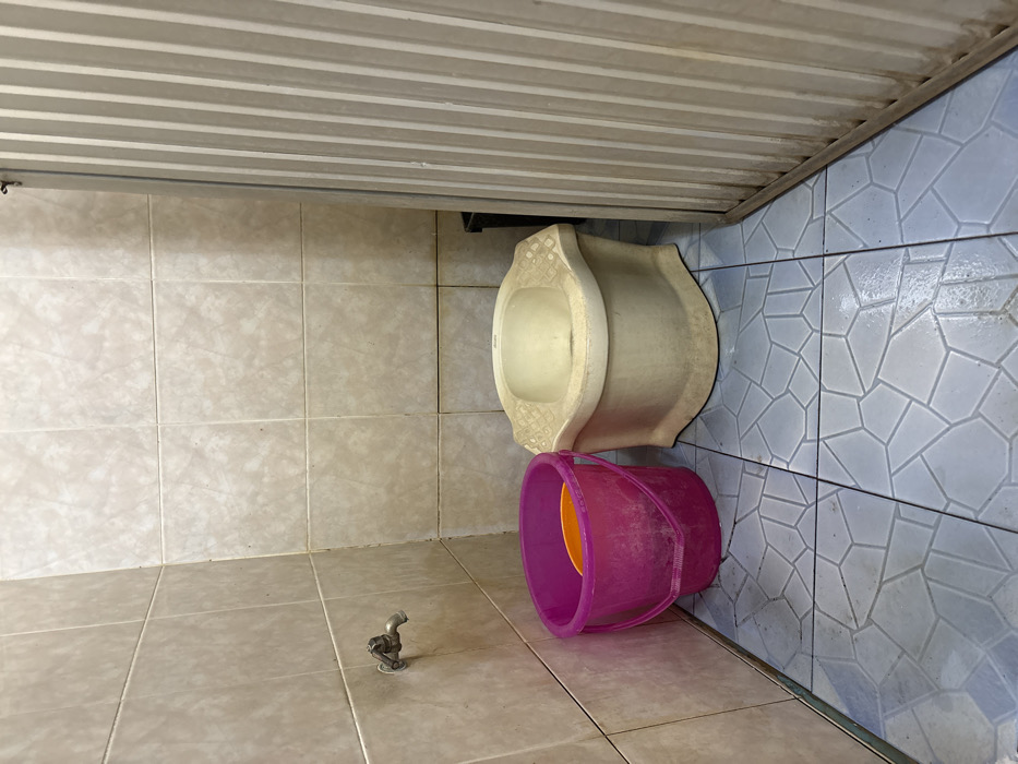 The scene depicts an interior view of a restroom facility located in Ban Hin Sam Kon, Thailand. The walls are surfaced with light beige square ceramic tiles. The floor is tiled with light blue polygonal-patterned ceramic tiles, showing some wetness. A metallic spigot is affixed to the left wall. On the floor below the spigot, a magenta plastic bucket containing an orange plastic bowl is present. To the right of the bucket, an off-white ceramic squat toilet is positioned. A corrugated metal partition, light in color, is visible on the far right, partially obscuring a dark container behind it. No individuals are present.