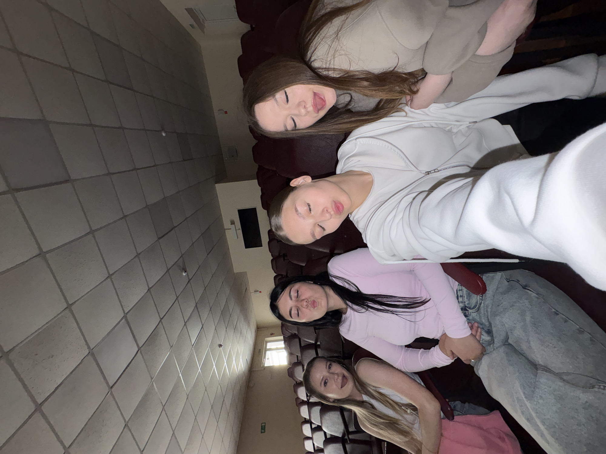 Four young women are seated in a row of dark red theater or auditorium seats. The room has a dropped ceiling with square acoustic tiles and fluorescent lighting. The walls are a neutral color, and there is a window with light coming through on the left side of the frame. In the background, more rows of seats are visible. One woman on the far left has blonde hair and is wearing a white tank top and a pink skirt. The woman next to her has long black hair and is wearing a pink long-sleeved shirt and light blue jeans. The third woman from the left has her hair pulled back and is wearing a white zip-up hoodie. The woman on the far right has long brown hair and is wearing a beige hooded sweatshirt. All four women appear to be looking towards the camera, with some making playful expressions. One woman is sticking her tongue out. The image appears to be taken in the city of Chaikovsky, Russia, likely in an educational or community building's auditorium.