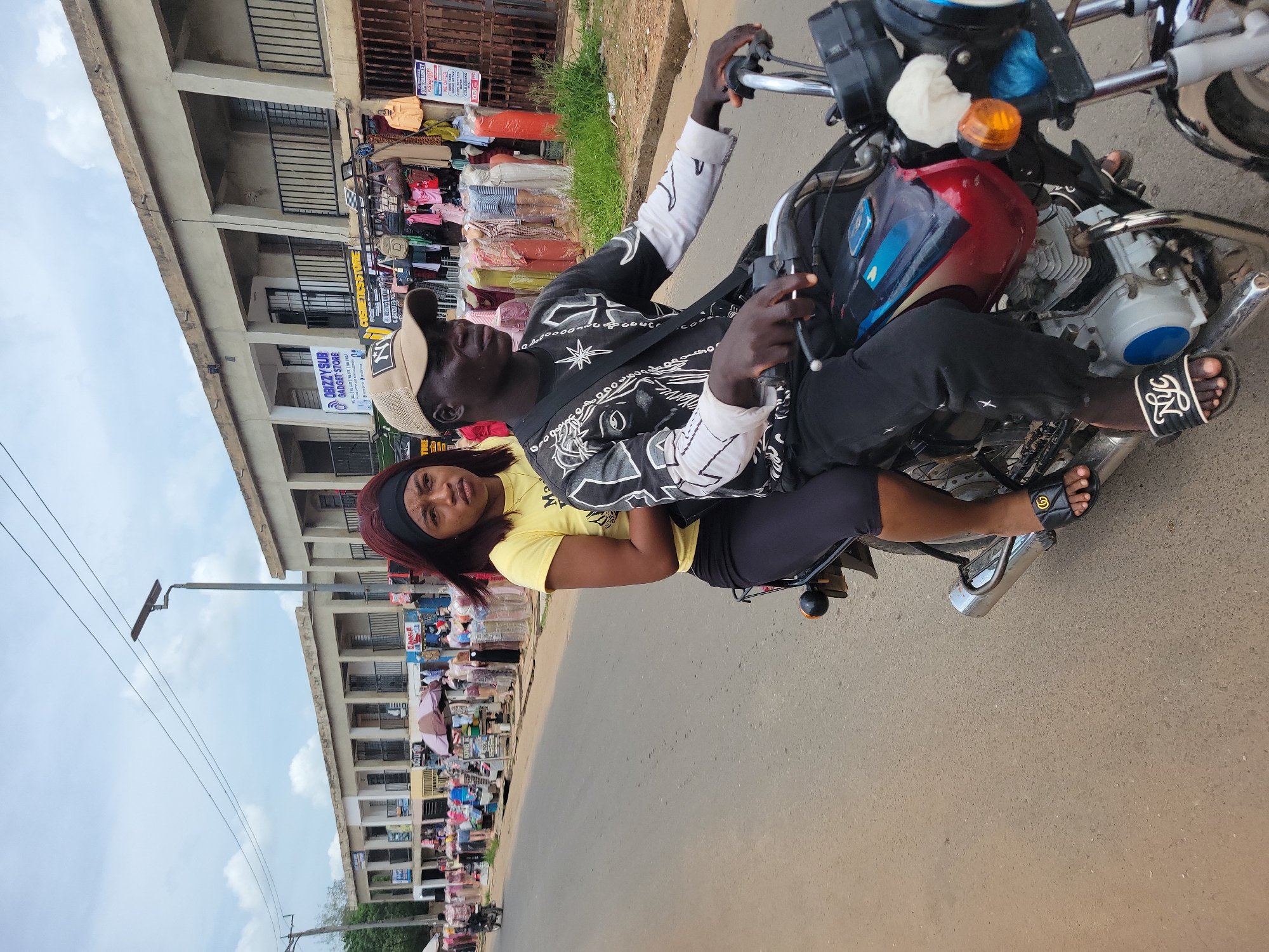 In Egbagu, Nigeria, a medium shot shows a man and a woman on a motorcycle. The man is wearing a dark cap, a black and white patterned jacket, and black pants. He is driving the motorcycle. The woman, with a black headband and wearing a yellow shirt and black pants, is seated behind him, holding onto him. Both are wearing open-toe sandals. The motorcycle is red and silver. The background features a multi-story building with shops and stalls selling various goods. A street lamp post is visible to the left. The road is paved. One of the shop signs reads "OBIZZY SUB GADGET STORE".