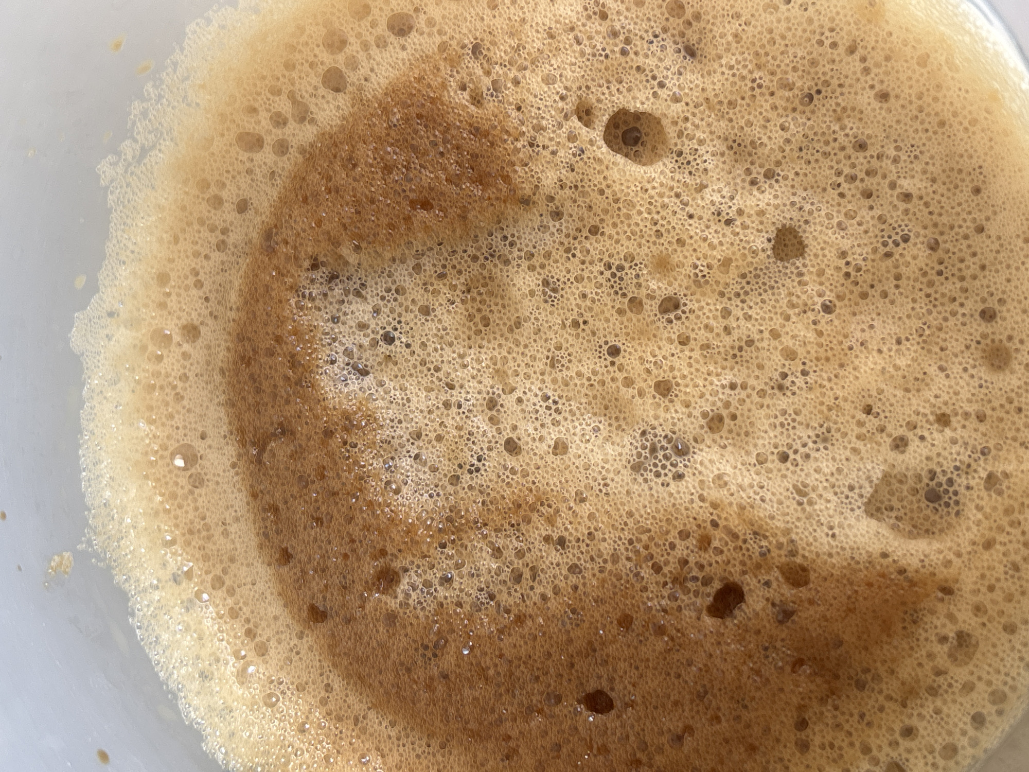 An extreme close-up shot captures the intricate surface of a frothy, brown beverage within a light-colored container, possibly a cup or mug. The primary subject is the rich, textured foam, composed of countless bubbles ranging in size from minuscule to several millimeters across. The foam displays a spectrum of light to medium brown and beige tones, with darker brown areas visible beneath and swirling through the lighter froth. These darker patches create abstract, organic patterns within the bubbly surface. The light-colored rim of the container is partially visible on the left and bottom edges, suggesting a white or light grey interior. There are no people, explicit activities, or visible text in the image. The scene is a static depiction of the beverage's surface, with no discernible time of day, weather, or location cues directly inferable from the image itself, though the context provided indicates the location is Plovdiv, Bulgaria. The lighting is even, highlighting the three-dimensional quality of the foam and its varied bubble structure.