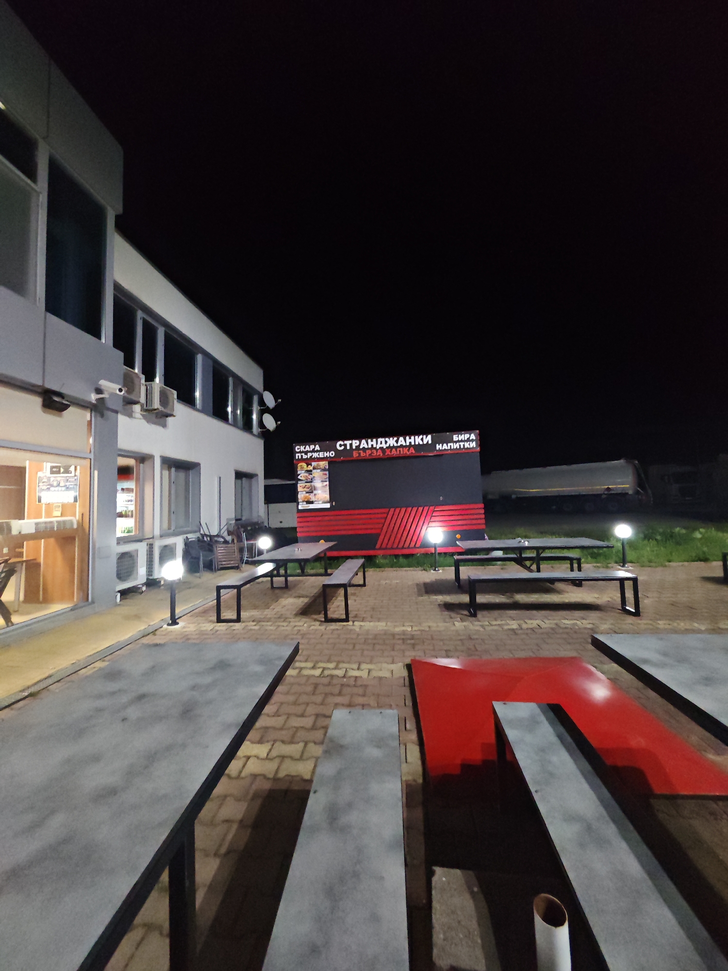 The image shows an outdoor dining area adjacent to a building at night. The building is a light-colored, two-story structure with several windows and air conditioning units mounted on the exterior. A brightly lit doorway is visible on the first floor.
The dining area consists of several picnic-style tables with attached benches. The tables have gray tabletops, and one table has a section painted bright red. The ground is paved with reddish-brown bricks.
Behind the tables is a large sign with black and red horizontal stripes. There is text in a Cyrillic alphabet on the sign. Additional text is printed on top of the sign, "СКАРА, СТРАНДЖАНКИ, БИРА, НАПИТКИ".
Two white lights illuminate the sign and surrounding tables. A large truck or tanker can be seen in the background. The environment is dark, indicating it is nighttime. This area appears to be a rest stop or dining area associated with the building.
The image was taken in Yabalchevo, Bulgaria.