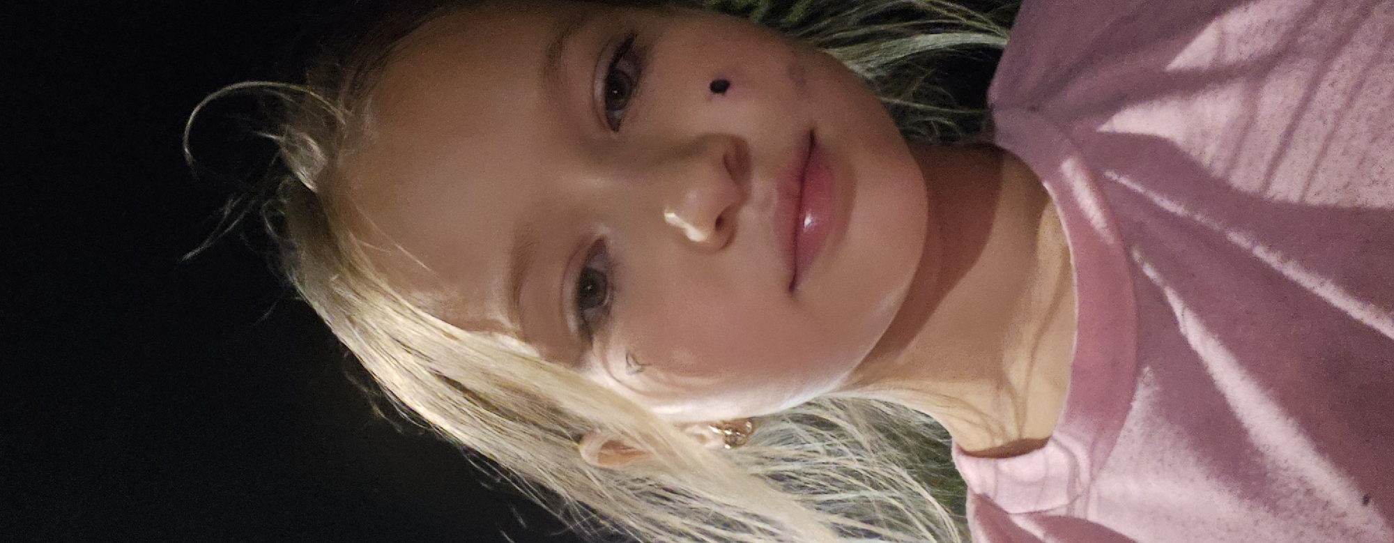 A close-up selfie of a young Caucasian girl with blonde hair, wearing a pink t-shirt, is taken outdoors at night. She has a small black dot painted on her cheek and a faint black marking under her left eye, resembling a tear or a beauty mark. Her hair is slightly windswept, and she is wearing a gold hoop earring in her left ear. The background is dark and indistinct, suggesting a nighttime setting. The image was captured in the city of Semey, Kazakhstan.