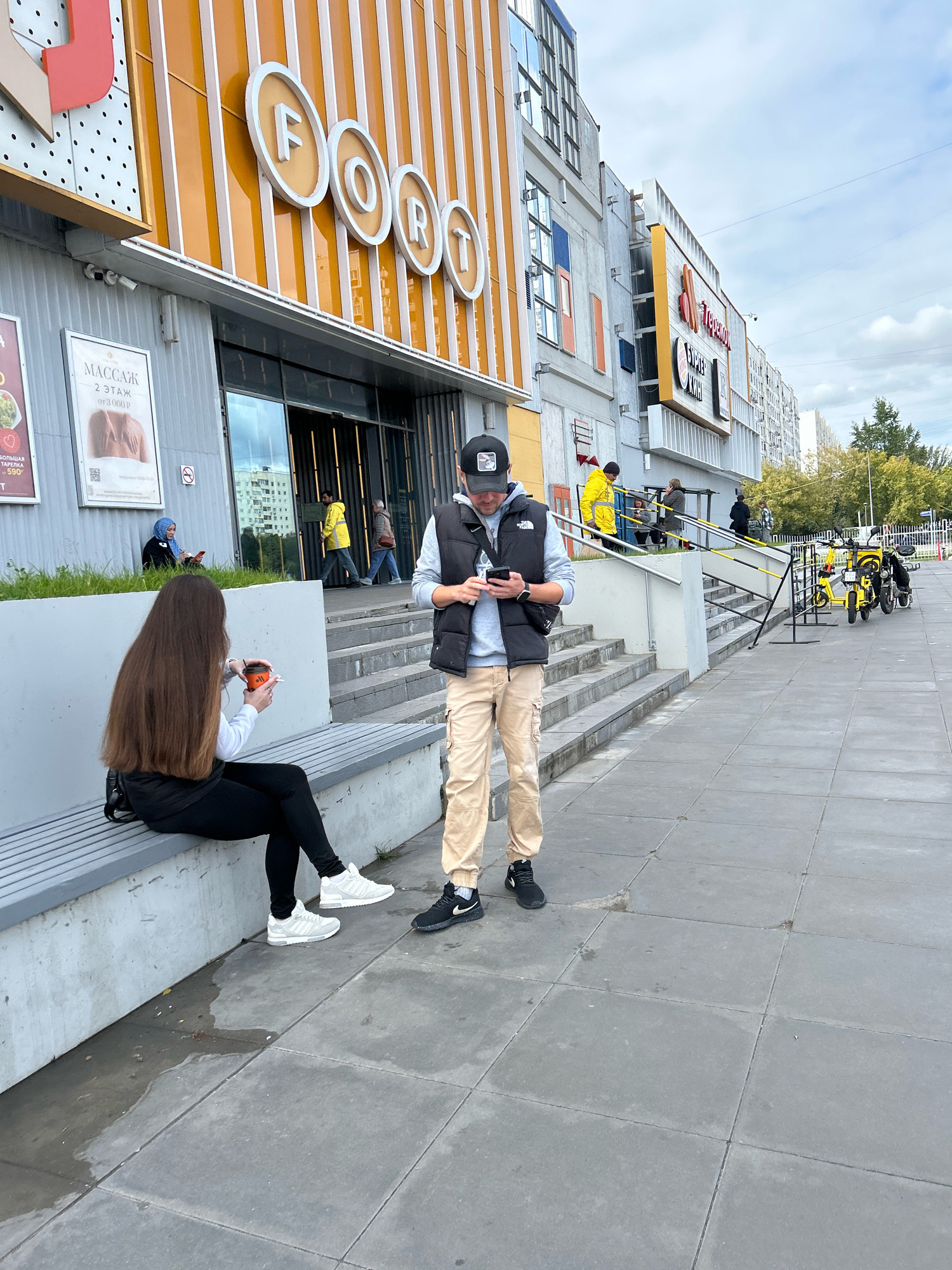 The image shows the exterior of a shopping center with a prominent "FORO" logo. Two young adults are the primary subjects. A young woman with long brown hair sits on a bench to the left, holding a cup and a cigarette. A young man stands in front of her, looking at his phone. Other people are visible in the background, including a person in a yellow jacket near the entrance of the shopping center. Several yellow electric scooters are parked to the right of the stairs. The pavement is made of large square tiles. The sky is overcast. The scene is likely in Moscow, Russia, given the Cyrillic signage on the shopping center.