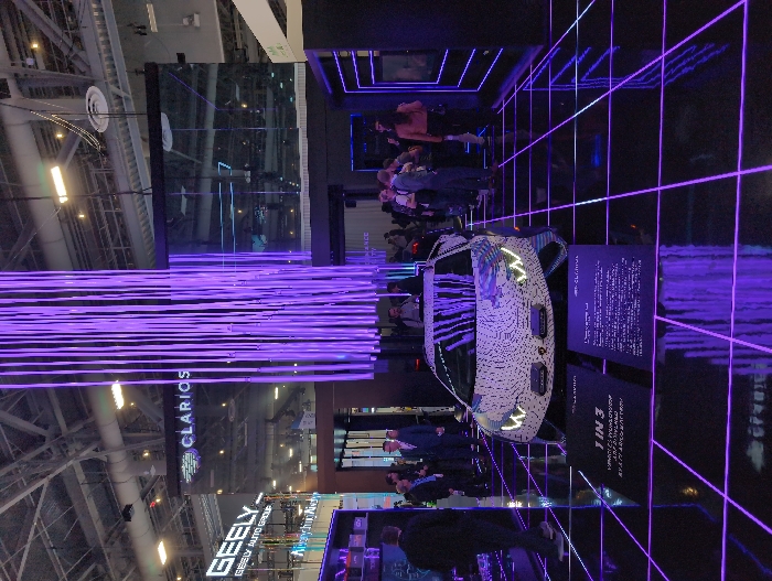 A white car is on display amidst an exhibition stand, its headlights illuminated. The backdrop features a neon grid pattern. People are gathered around the vehicle. The stand has a digital display with information on the car. Above the display, there are visible brands, "GEELY AUTO GROUP" and "CLARIOS." The exhibition is in a large venue with a high ceiling supported by metal beams.