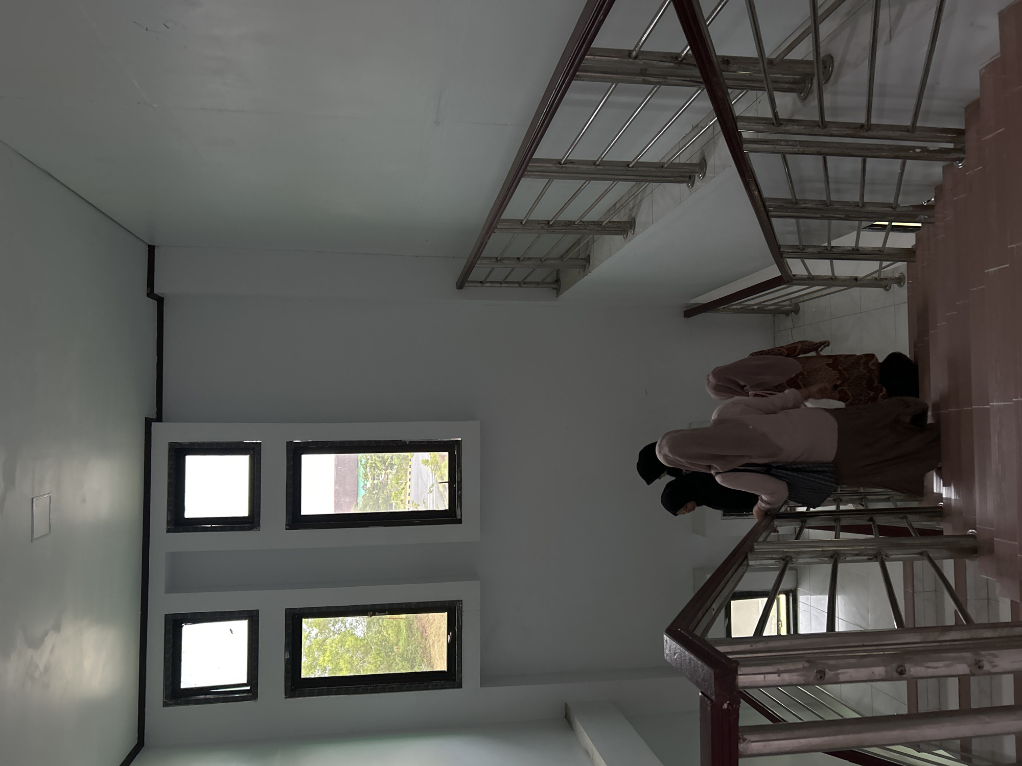 The image depicts an indoor stairwell in what appears to be a multi-story building, likely in Reuleut Barat, Indonesia, given the provided context. The perspective is from the bottom of the stairs looking up.
Three women are ascending the staircase. The woman in the foreground wears a light pink long-sleeved top, a striped grey and black garment over it, a long brown skirt, and a black hijab. Behind her, another woman is partially visible, wearing a brown patterned garment and a black hijab. A third woman, mostly obscured, wears a black top and black hijab. Their attire suggests traditional wear common in the region.
The stairwell features a staircase with dark reddish-brown wooden handrails and stainless steel balusters, leading up to a tiled landing. The walls are painted white. On the left wall, four small, rectangular windows are arranged in two rows of two, providing natural light and a view of green foliage outside, indicating it is daytime. A rectangular panel, possibly an access hatch or electrical panel, is also visible on the white wall to the left of the windows. The ceiling is white, and the overall lighting is bright and even. There is no visible text in the image.