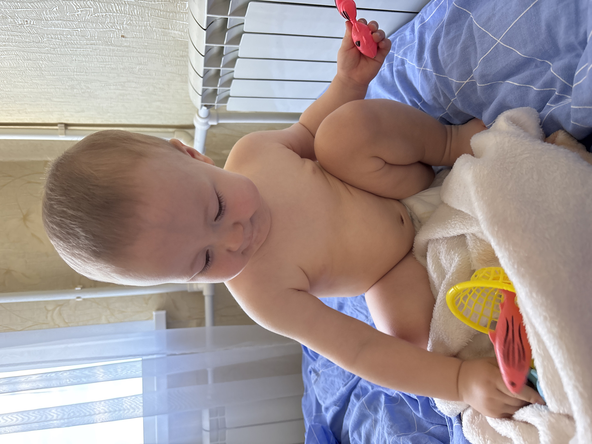 A baby is sitting on a blue blanket, covered by a white towel. The baby is wearing no clothes and is holding a pink toy fish in their right hand and a yellow fishing net in their left hand. The baby appears to be looking down at the toys. The scene is indoors, with a radiator and a window visible in the background. The window has white blinds. The lighting suggests it is daytime. The baby's expression is neutral. The location is Naberezhnye Chelny, Russia. There is no visible text in the image.