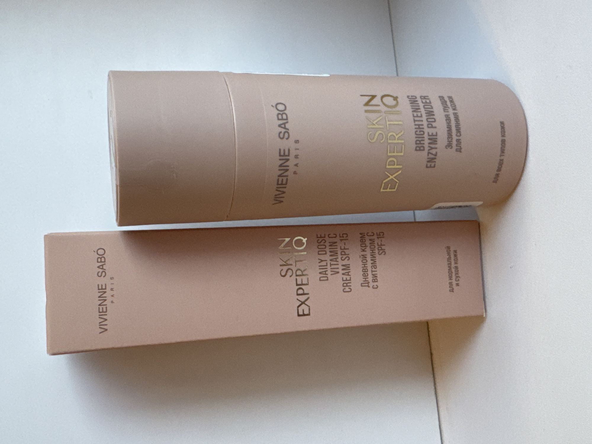 The image displays two VIVIENNE SABÓ branded cosmetic products, a box for "SKIN EXPERTIQ DAILY DOSE VITAMIN C CREAM SPF-15" and a cylindrical container labeled "SKIN EXPERTIQ BRIGHTENING ENZYME POWDER." The box is positioned to the left and slightly behind the powder container. Both items are a light peachy-pink color with gold lettering. The background appears to be a light-colored, textured surface, possibly a table or shelf, with a white wall behind. There are no people, animals, or any other distinguishing environmental features visible in the image. The lighting suggests an indoor setting with soft, diffused light. The location is not discernible from the image itself; information about Яромінскі сельскі Савет, Belarus, is external to the visual content.