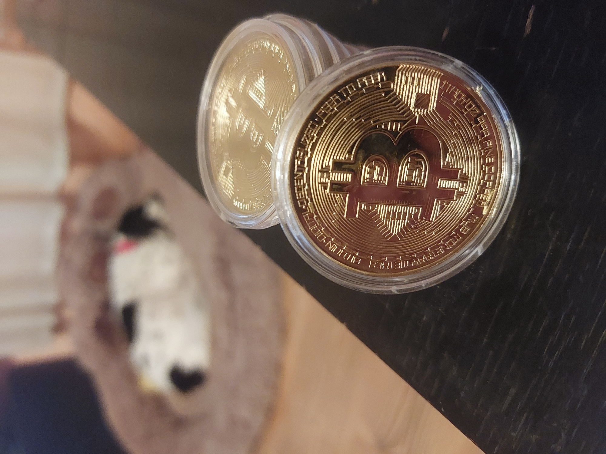 The image shows two gold-colored Bitcoin commemorative coins, encased in clear plastic protectors, sitting on a dark, textured surface that appears to be a wooden table. One coin is slightly in front of and to the right of the other, partially overlapping it. The coins feature the Bitcoin logo and intricate designs. The background is blurry and appears to show a soft, textured object, perhaps a stuffed animal or part of a piece of furniture. There is no information in the image to indicate that this image was taken in Tsarevo, Bulgaria.