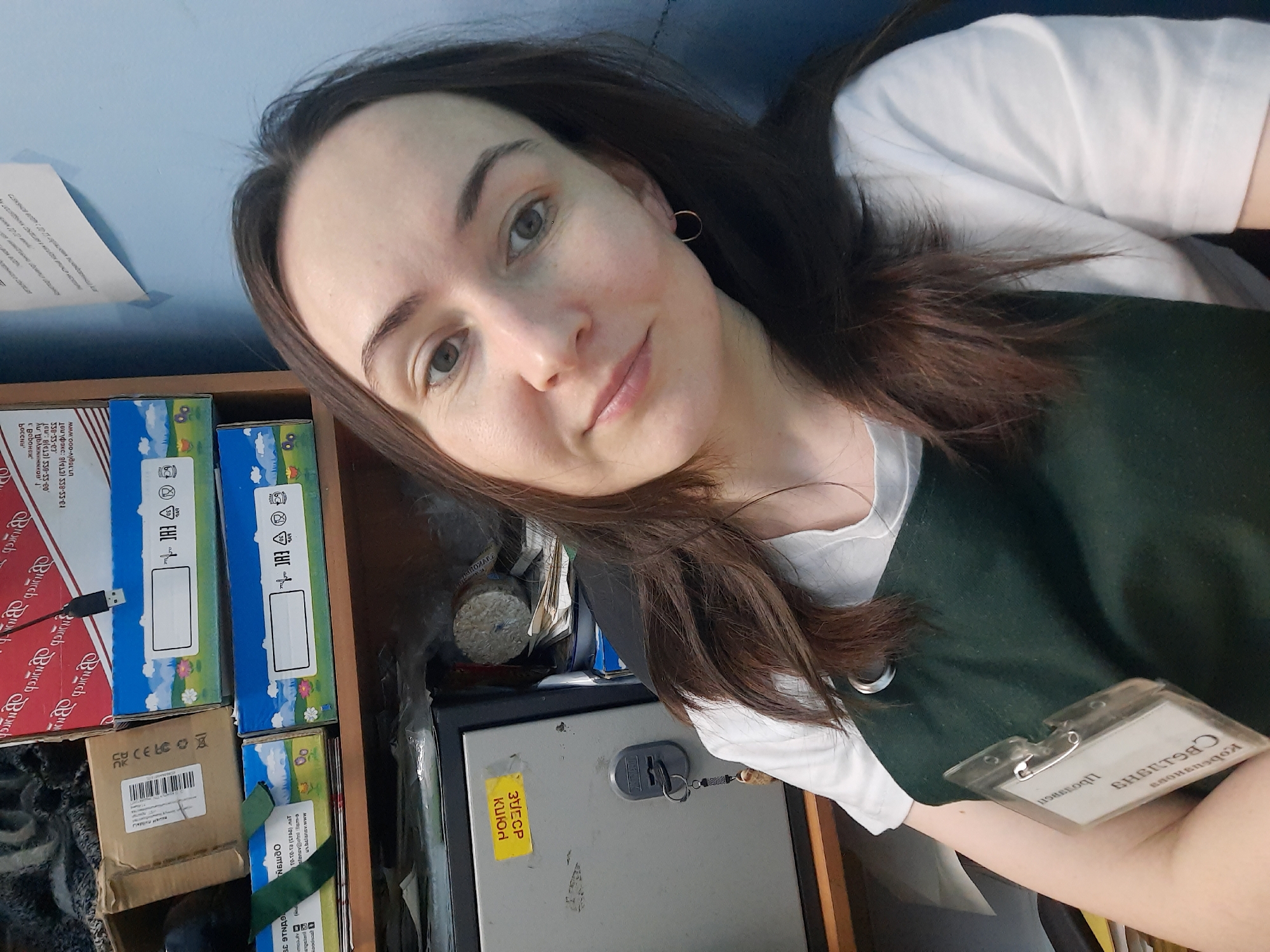 A selfie taken by a woman with long brown hair. She has fair skin and is wearing a white t-shirt under a dark green garment. She is indoors, as evidenced by the presence of furniture. To the left is a wooden shelf with boxes of different sizes and designs. Behind her is a light blue wall with a piece of paper attached. A metal safe with a key is also visible. She is wearing a name tag with the name "Kobaenous." Some of the boxes on the shelves contain the words "Burch." The Cyrillic text on one box includes information such as phone numbers, and a website. Another box includes a green tag that reads "Belangev" and "Krasnogorskoye." A yellow sticky note on the safe has the words "31ECP Kuion."