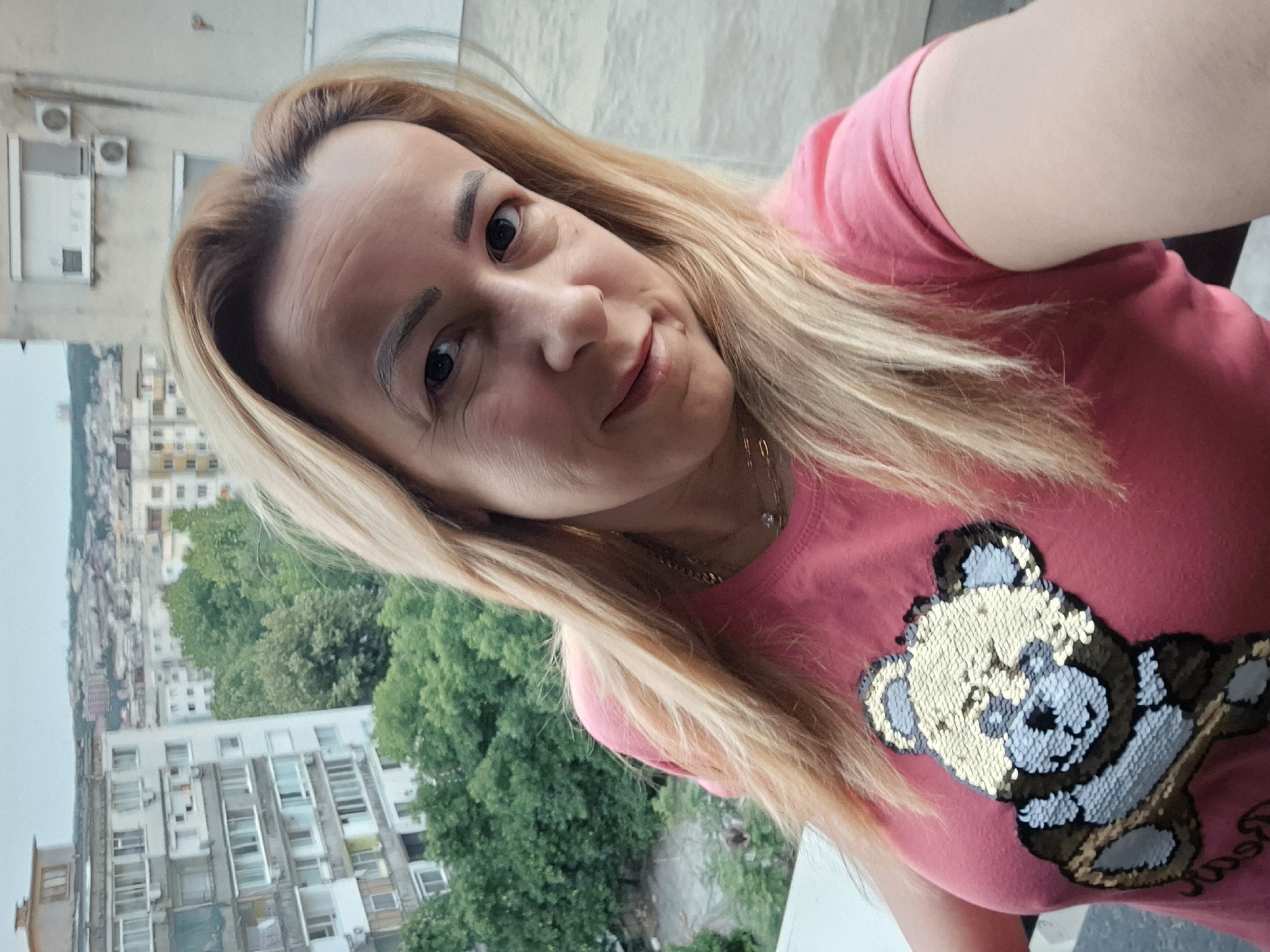 In a selfie taken from a balcony, a Caucasian woman with blonde hair and fair skin is pictured from the chest up. She is wearing a pink t-shirt with a sequined teddy bear design. In the background, an urban landscape unfolds with apartment buildings and trees under an overcast sky. The location is identified as Unknown, Unknown.