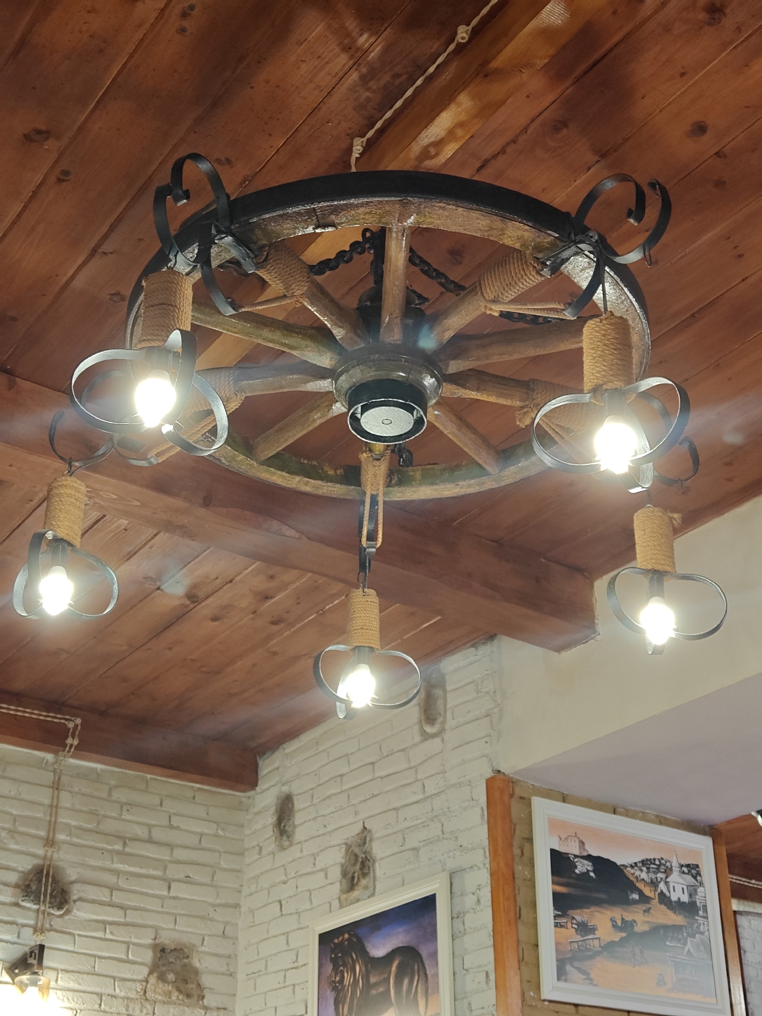 A rustic chandelier, crafted from a large wooden wagon wheel, dominates the center of the image, suspended from a warm-toned wooden paneled ceiling. The wheel features a dark metal band around its rim and spokes radiating from a central hub. Six light fixtures hang from the wheel, each encased in black, decorative metal loops and suspended by a rope-wrapped cylindrical element. The bulbs emit a bright, clear light, illuminating the space below.
The ceiling above is constructed from wooden planks, running parallel to each other, with visible grain and natural imperfections. A thin, light-colored rope or wire is subtly visible along the ceiling, near the top right.
Below the chandelier, a white brick wall, interspersed with irregular natural stone inserts, forms part of the room's interior. Two framed paintings adorn the wall. The one on the right, predominantly in shades of gray and white with orange-brown sky accents, depicts a town or village scene with buildings clustered around a hillside crowned by a structure, possibly a castle or church. The painting on the left, partially visible, appears to show an animal, possibly an elephant or another large creature, with a darker color scheme.
The scene suggests an indoor setting, likely a restaurant, tavern, or a room designed with a traditional or rustic aesthetic, possibly in Tbilisi, Georgia, where such decor is common. The bright artificial lighting indicates it might be evening or an interior that relies on artificial illumination. No people are visible, and there is no discernible text.