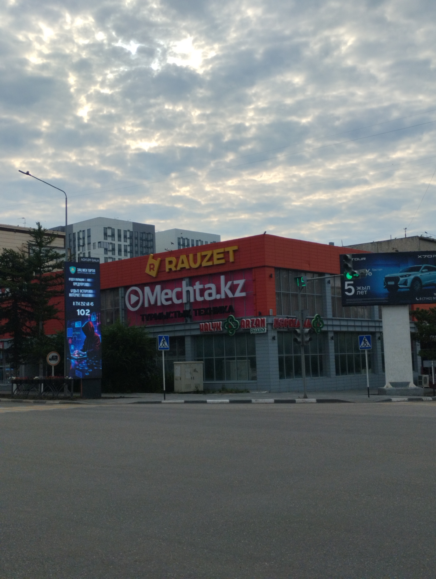 The image shows a street view of a commercial building complex in Kostanay, Kazakhstan. The main building in the foreground is a bright orange structure with the text "RAUZET" prominently displayed in large red letters. Below "RAUZET", the text "Mechta.kz" is visible in large white letters, with "ТУРМЫСТЫК ТЕХНИКА" (Household Appliances) written underneath. To the left of the main building, a tall digital billboard displays various information, including the number "102" and an image of a person with a hoodie using a laptop.
The building also features a sign that reads "ARZAN PHARM". A traffic light is visible, showing a green light. A large billboard advertising a car with the text "X70 PRO" and "5 ЖЫЛ ЛЕТ" (5 Years) is on the right side of the image.
In the background, modern-looking multi-story buildings are visible. The sky is overcast with clouds, and the lighting suggests it is daytime, possibly late afternoon or early morning. The street in the foreground is paved asphalt, with pedestrian crossings and street furniture like benches and trash cans. A street lamp is also visible. There are no people clearly visible in the image.