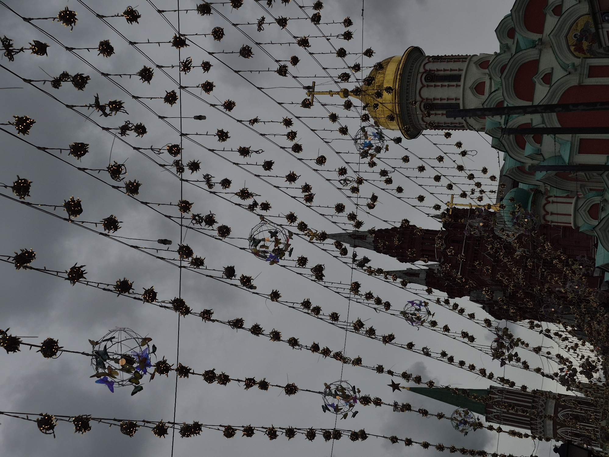 This is a low-angle, close-up, eye-level shot of a street decorated with strings of lights and spherical ornaments, many with spiky protrusions and small, glowing bulbs. The lights are arranged in diagonal lines against a backdrop of an overcast, gray sky. Interspersed among the lights are decorative spheres containing colorful butterfly-shaped ornaments. In the background, the tops of several historic buildings are visible. On the right, a building with a golden dome and a cross is prominent, along with adjacent structures featuring red, white, and teal architectural elements. To the left, a tall red building with ornate details and a green spire topped with a star can be seen. The overall impression is of a festive streetscape under a cloudy sky, likely in a historic urban center.
