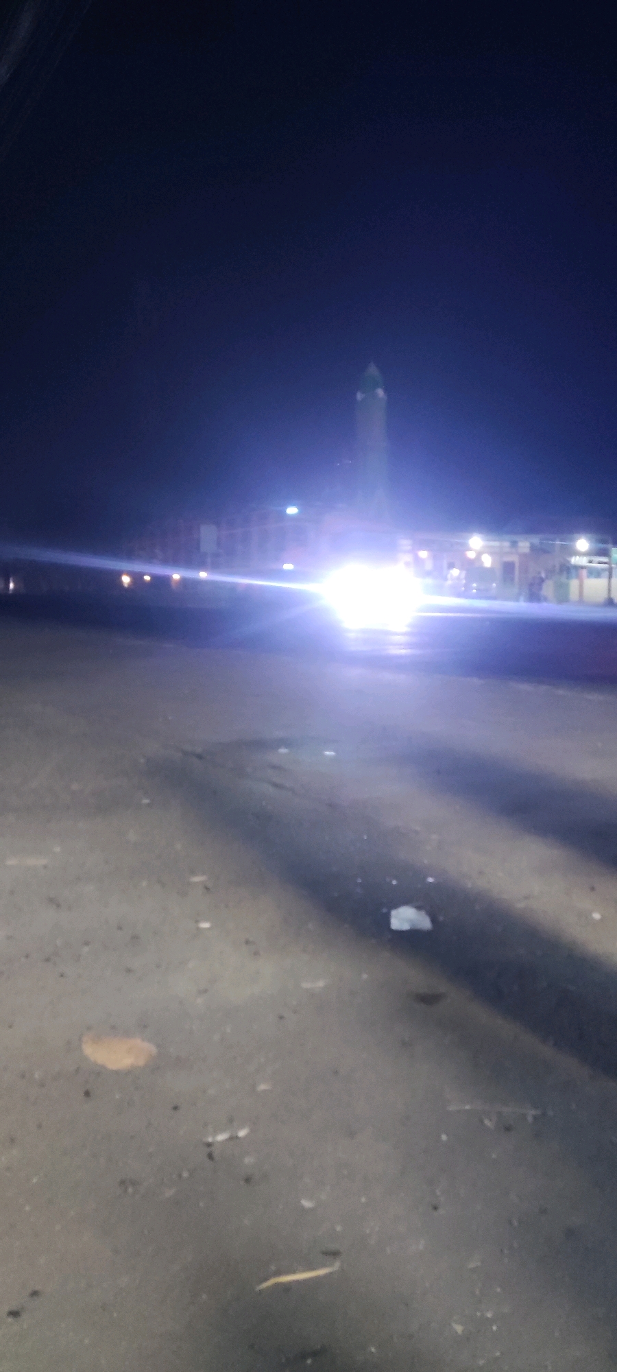 The image depicts a nighttime scene in Pelayangan, Indonesia. A tall, slender structure, likely a minaret, is illuminated by bright lights and stands prominently against the dark sky. The structure has a green cap. In front of it, a building with multiple windows is visible, also lit up. The foreground is a dark, paved area, possibly a street or a plaza, with tire tracks and scattered debris. The overall impression is one of a religious or public building at night, with strong artificial lighting creating a bright, somewhat hazy atmosphere.