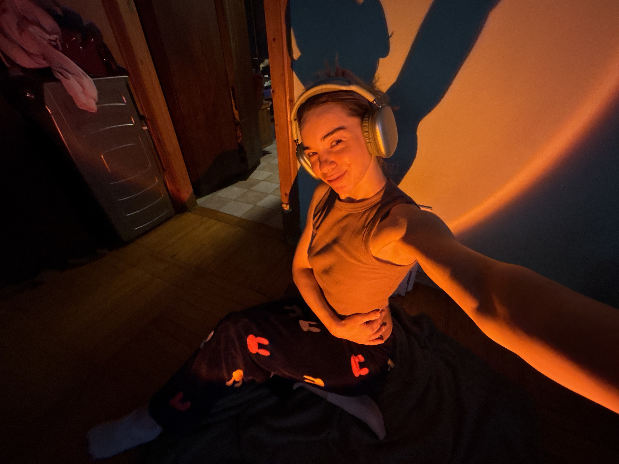 A young woman with headphones poses for a selfie in a dimly lit room. She wears a tank top and patterned pajamas, sitting on a dark blanket. A room divider and a cabinet are visible in the background. An orange light projects onto the wall behind her, creating a shadow. The location is Unknown, Russia.